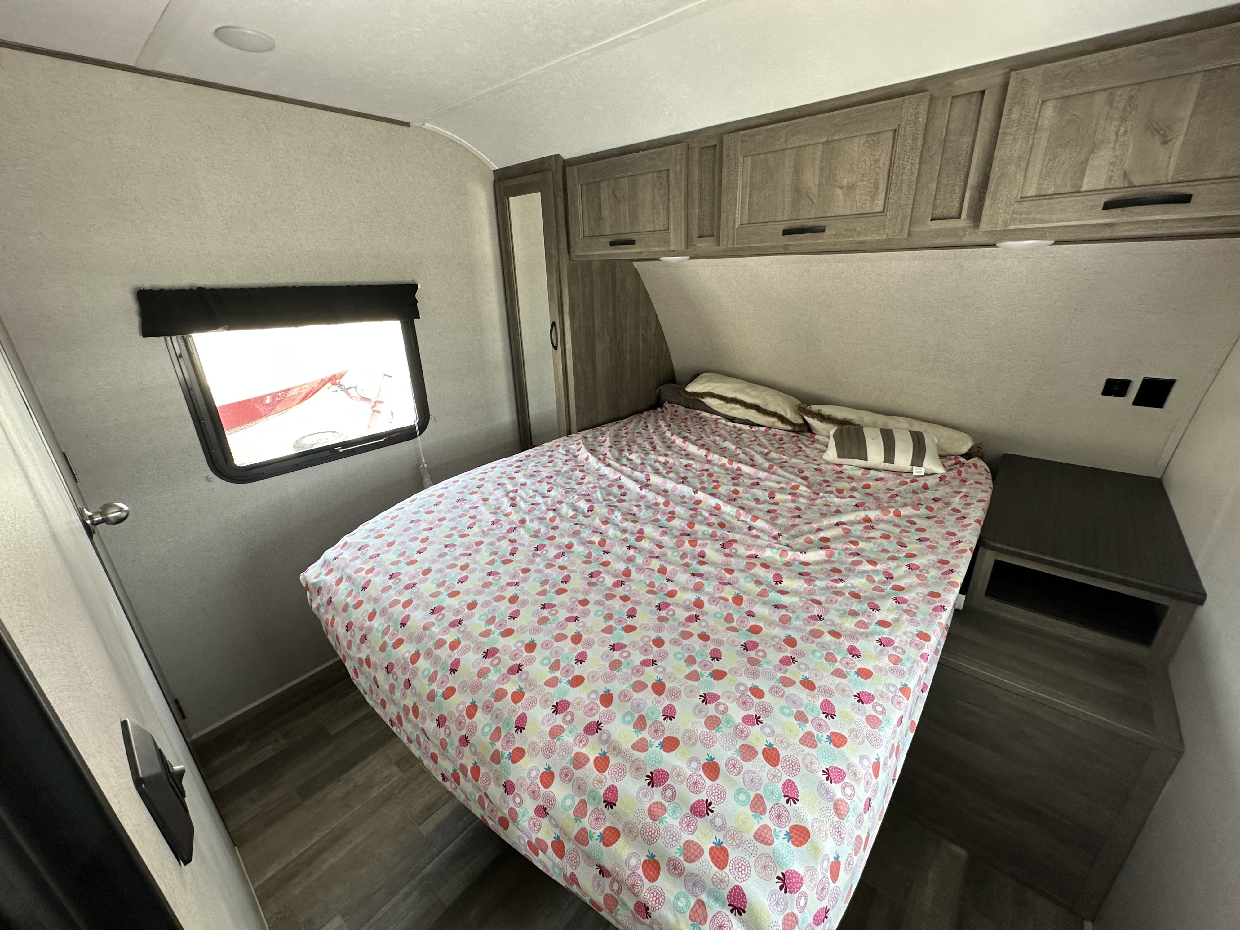 RV rental bedroom with patterned bedding, overhead storage, a small nightstand, and a window.. Forest River Vibe 2019