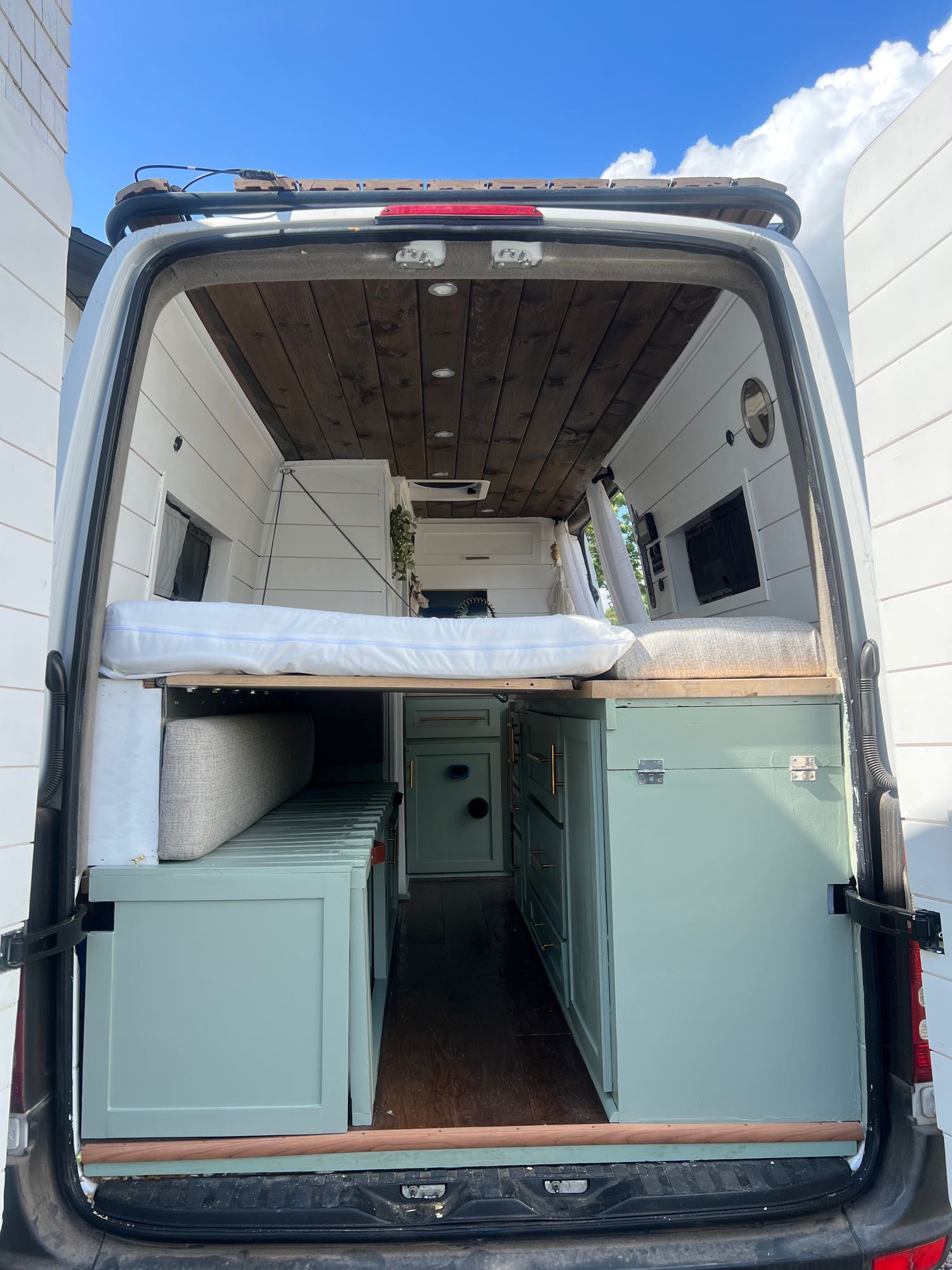 View into an RV rental van from its open back doors, showcasing the raised bed, wood ceiling, and light green cabinetry.. Mercedes Sprinter 2012