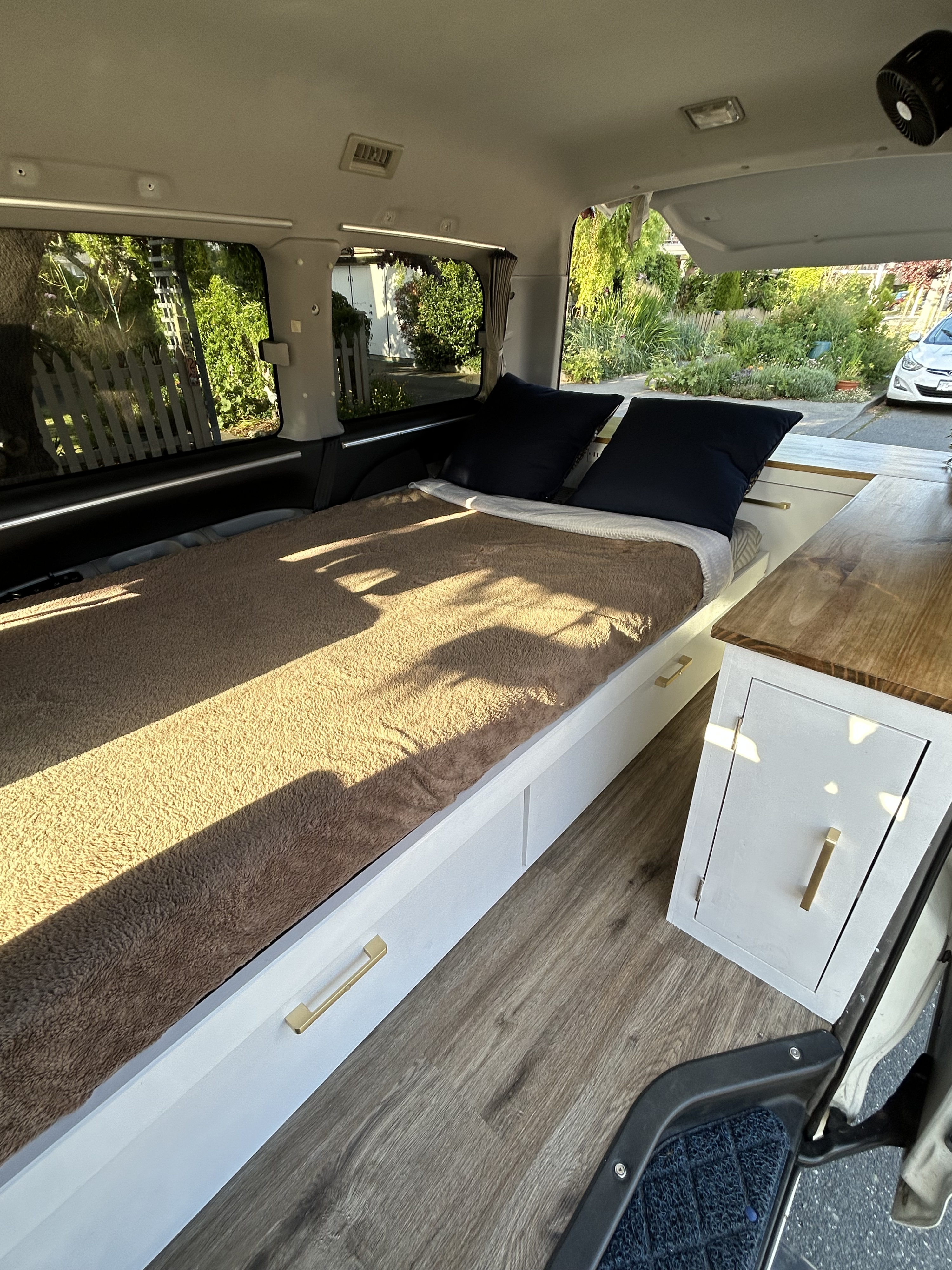 Cozy RV rental interior features a comfy bed with built-in storage, wooden accents, and sunlit windows, ready for adventure.. Mitsubishi Delica 1999