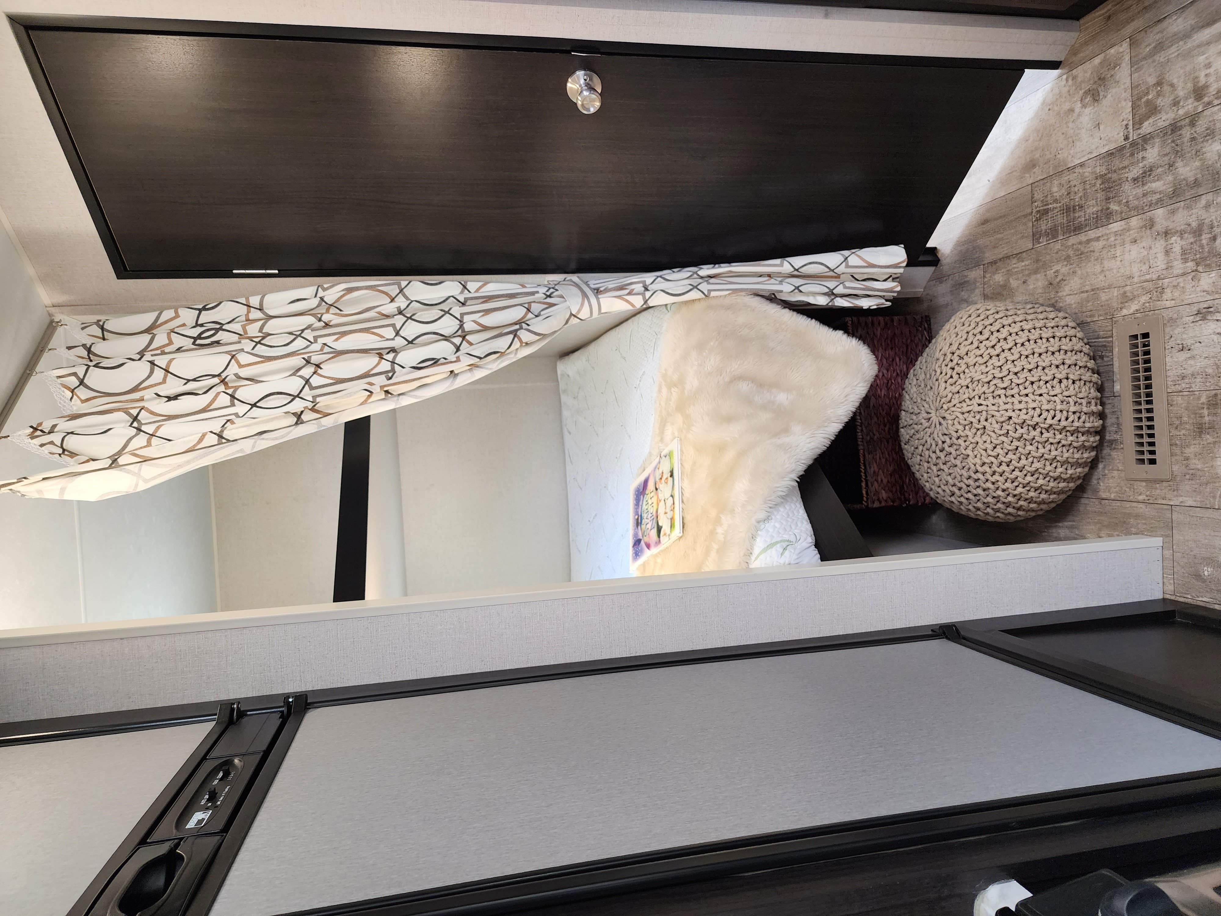 RV rental interior with a cozy bed, patterned curtain, dark wood upper cabinet, and a knitted pouf on wood-look floor.. Venture Sporttrek 2018