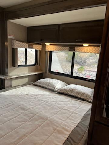 Cozy RV rental bedroom featuring a comfortable bed with pillows, patterned window valances, and ample overhead storage cabinets.. Thor Quantum 2020