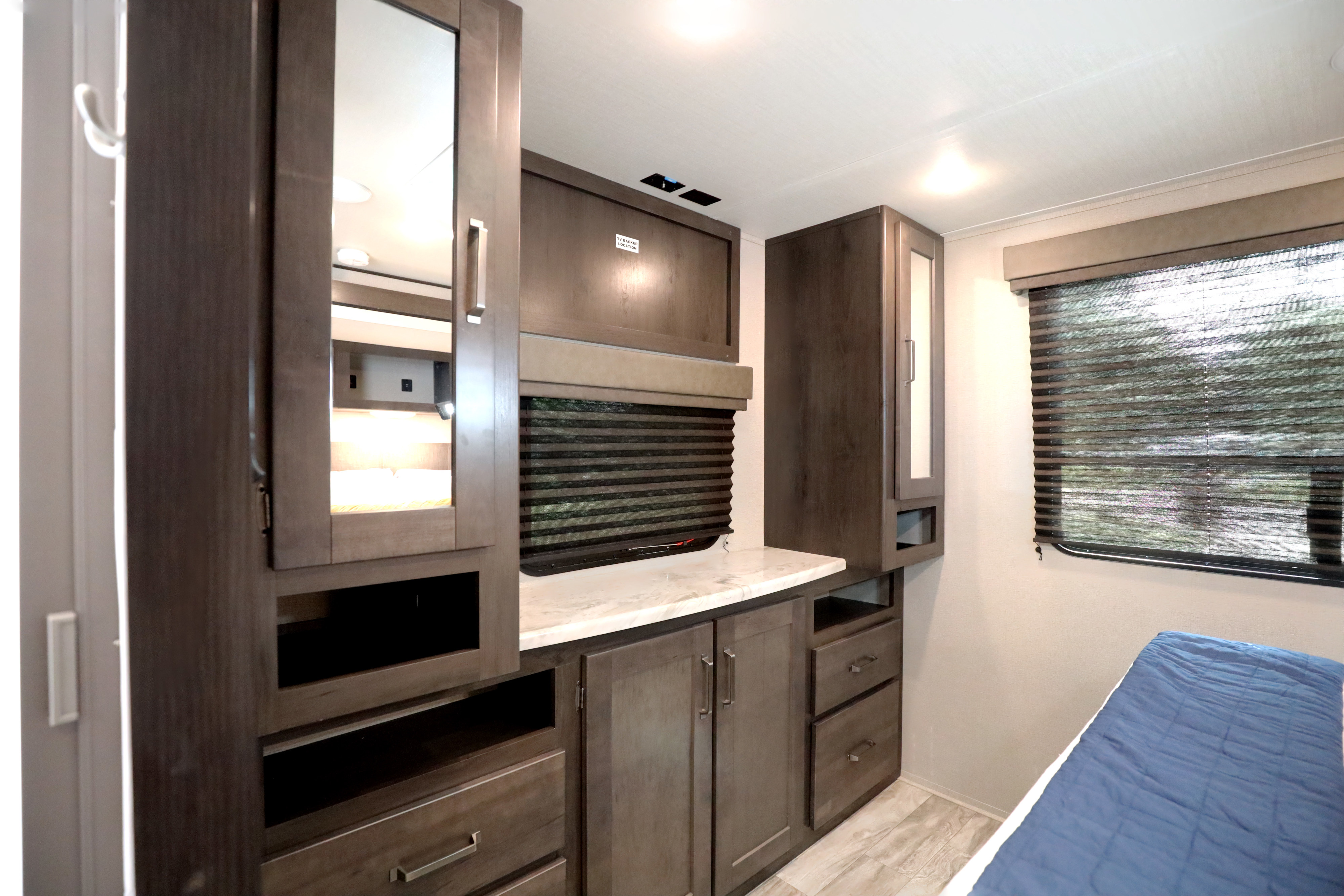 This RV rental bedroom offers dark wood cabinetry, a mirror, window with blinds, and a cozy bed.. Grand Design Transcend Xplor 2022