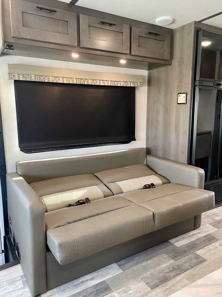 An RV rental's versatile living space: a beige convertible sofa with seatbelts, overhead storage, and window with shade.. Forest River FR3 32DS 2023