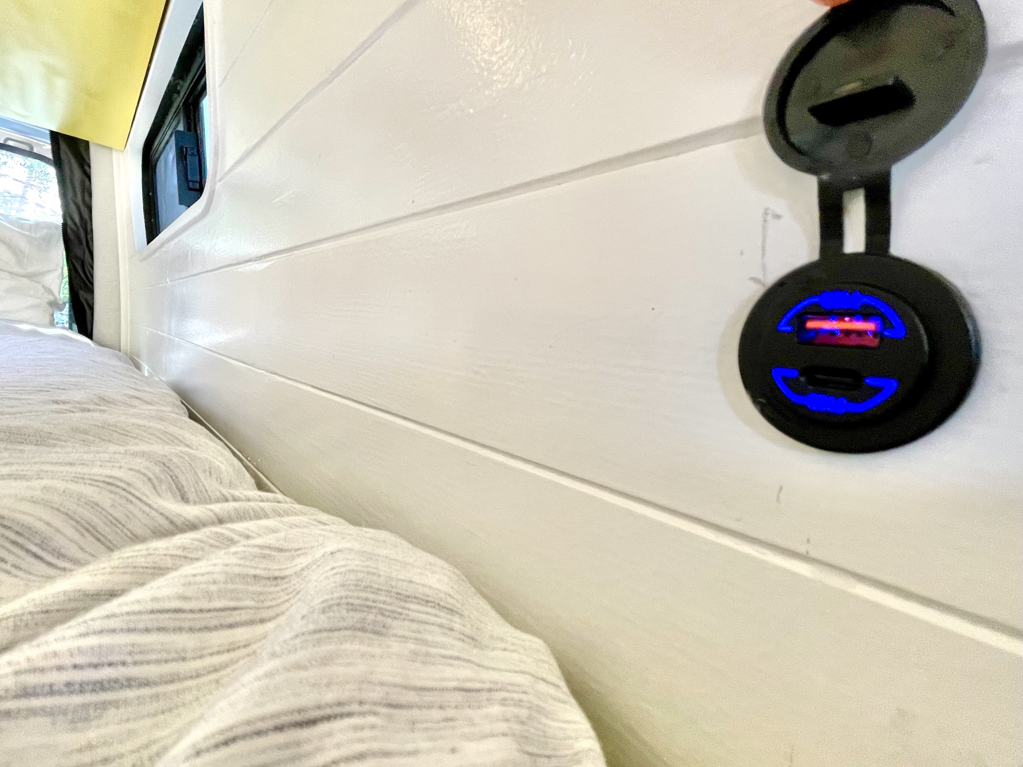 Inside an rv rental, a comfortable bed next to a white plank wall featuring a window and illuminated dual USB charger, perfect for device charging.. Mercedes-Benz sprinter 2020
