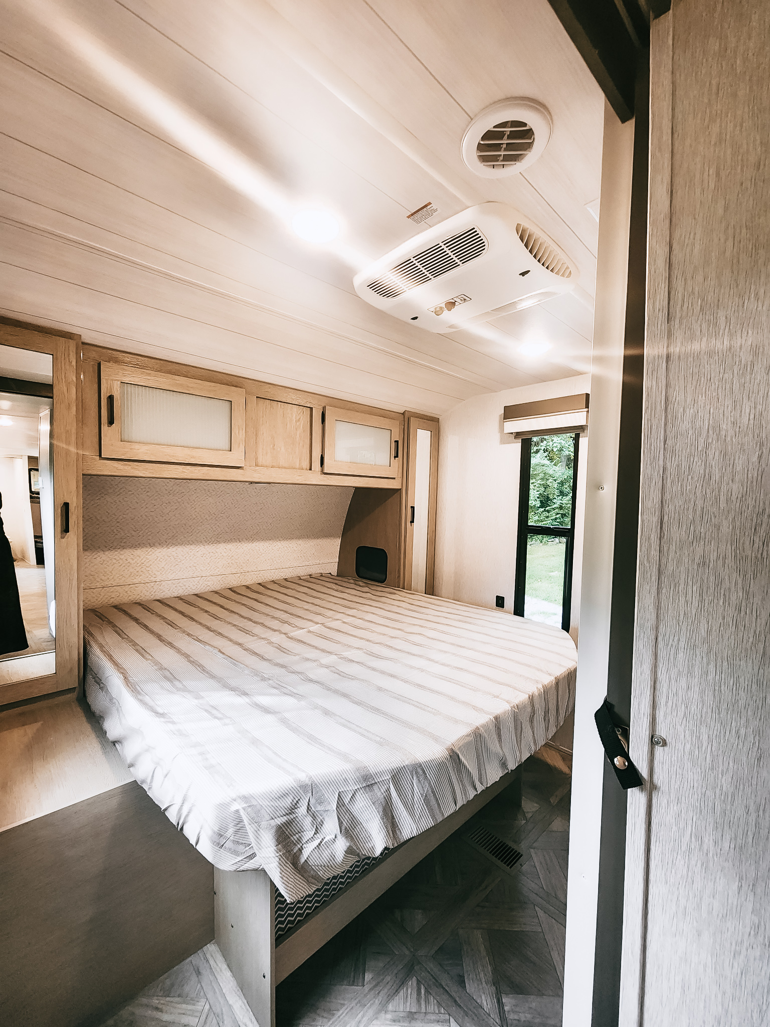 Inside a bright RV rental bedroom showing a striped bed, wood cabinets, ceiling AC, and a window view.. Forest River Wildwood 2023