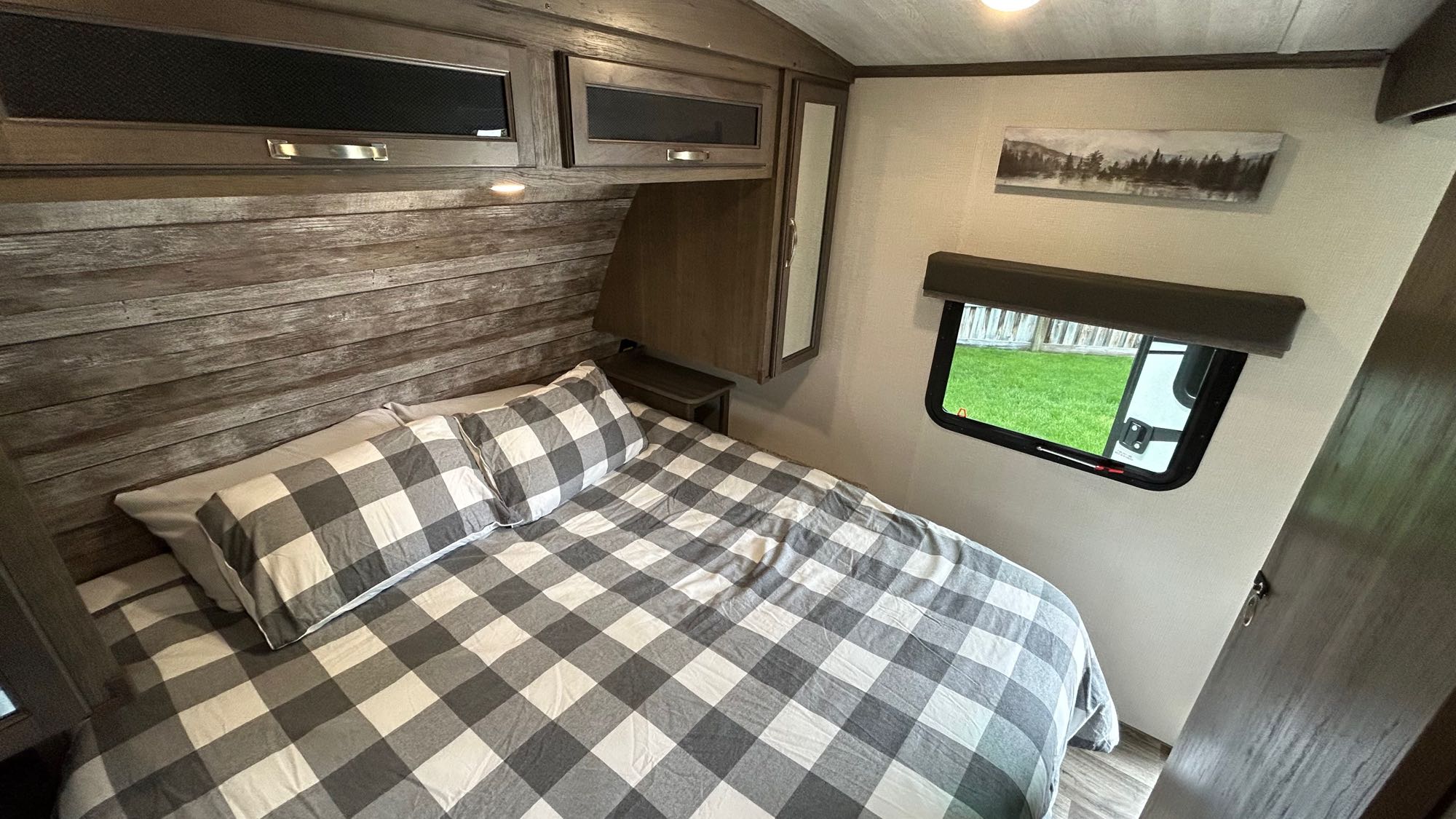 Inside an RV rental bedroom: a bed with gray buffalo check bedding, distressed wood wall, overhead cabinets, and side window.. Keystone Cougar Half-ton 2022
