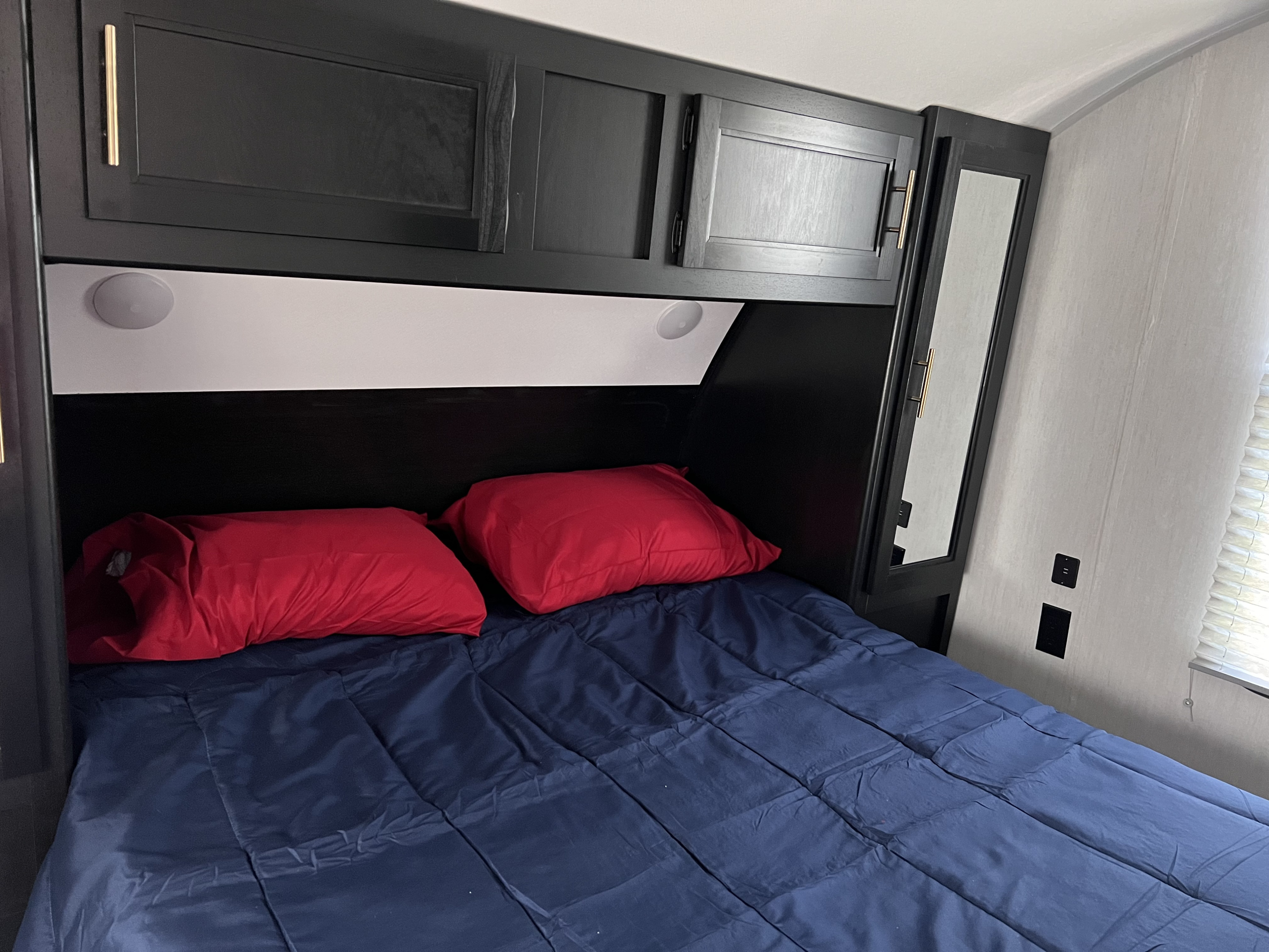 Inside an RV rental: a compact bedroom featuring a blue bed, red pillows, dark cabinets, and convenient built-in lighting.. Forest River Cherokee 2022