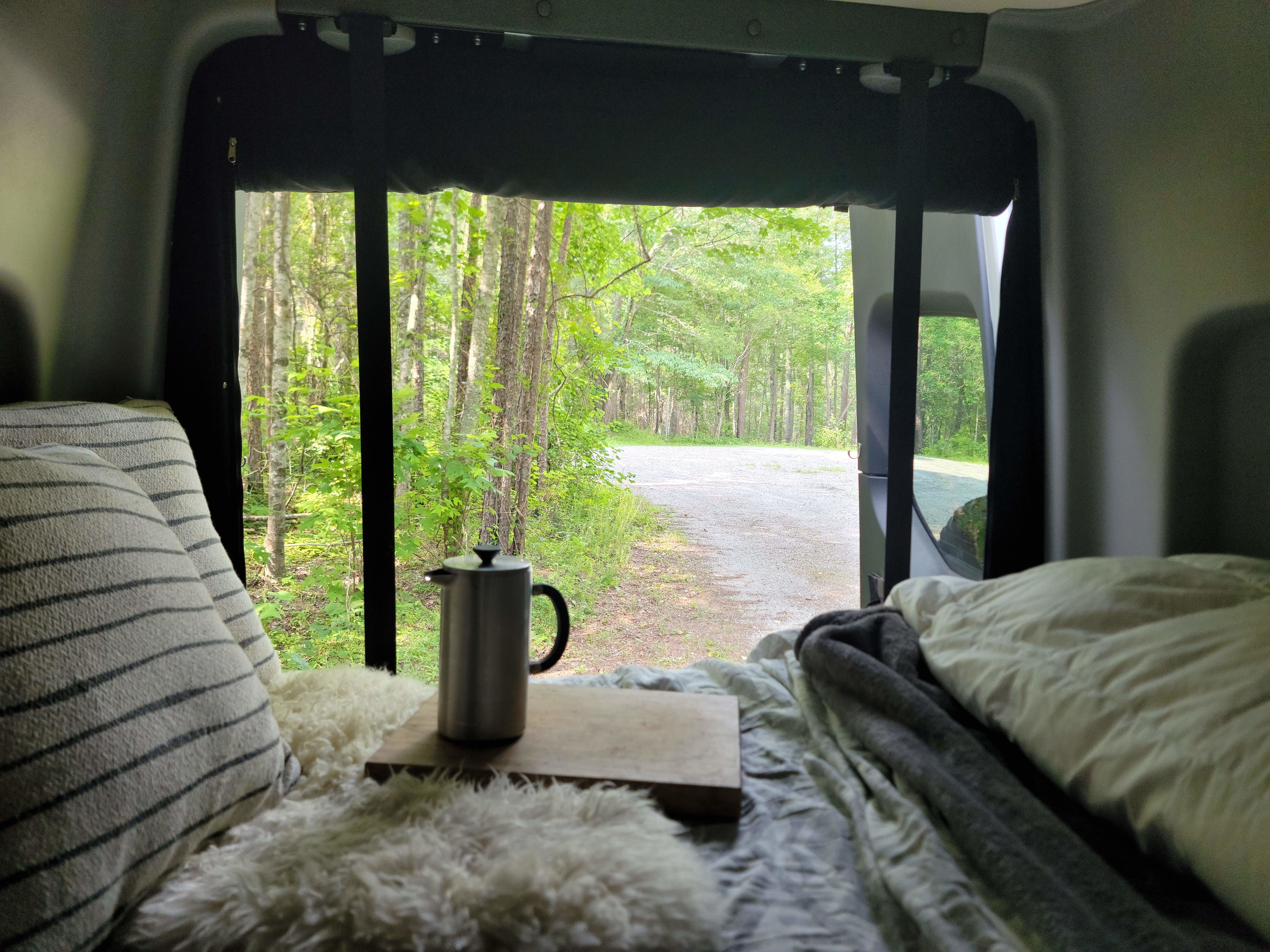 Cozy RV rental interior: coffee press on bed looking out to a forest road, surrounded by lush trees.. Winnebago Revel 2022