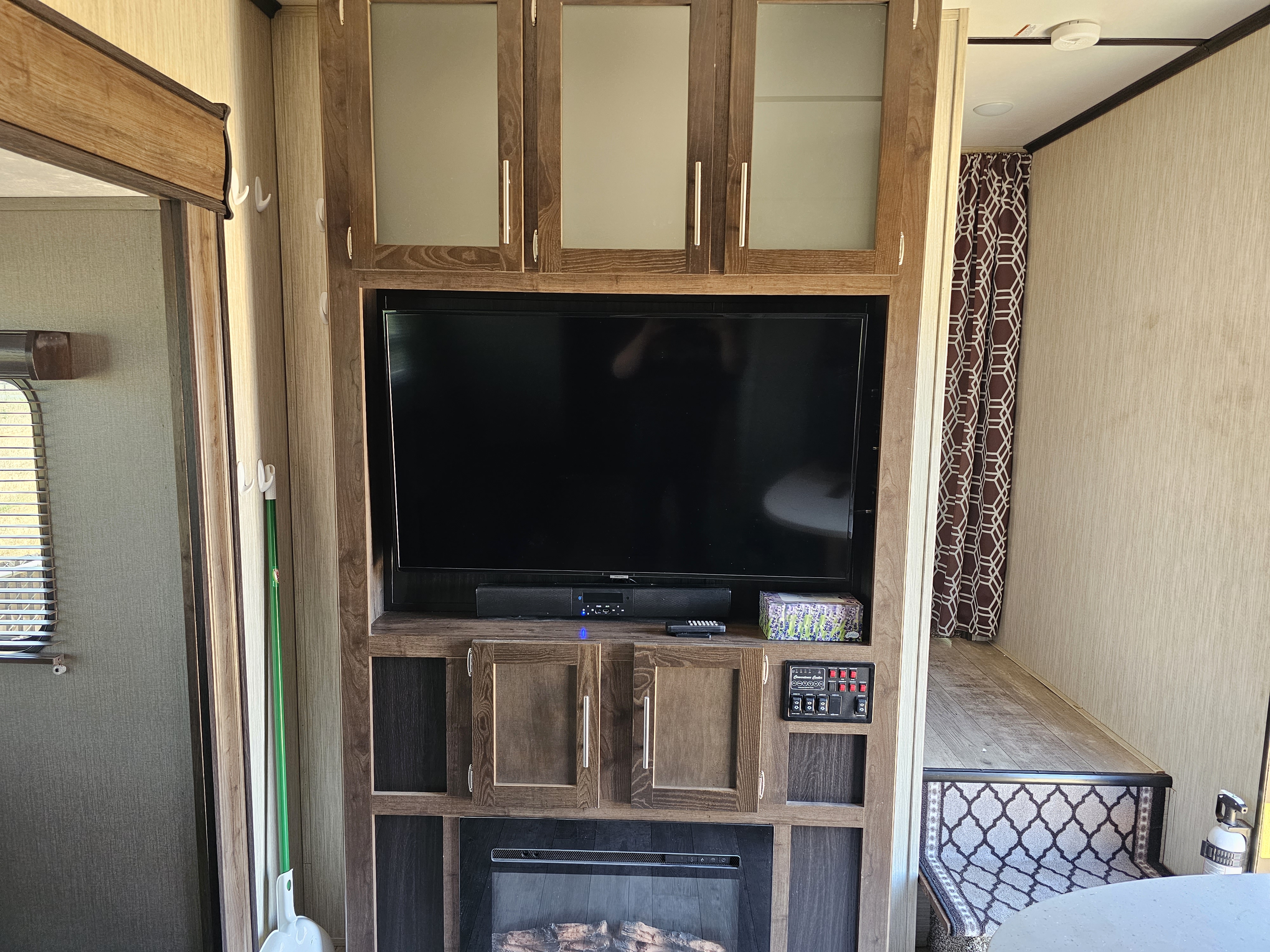 Inside this RV rental: a modern entertainment center with a TV and fireplace, plus essential RV controls, and a raised bed area.. Crossroads RV Volante 2019