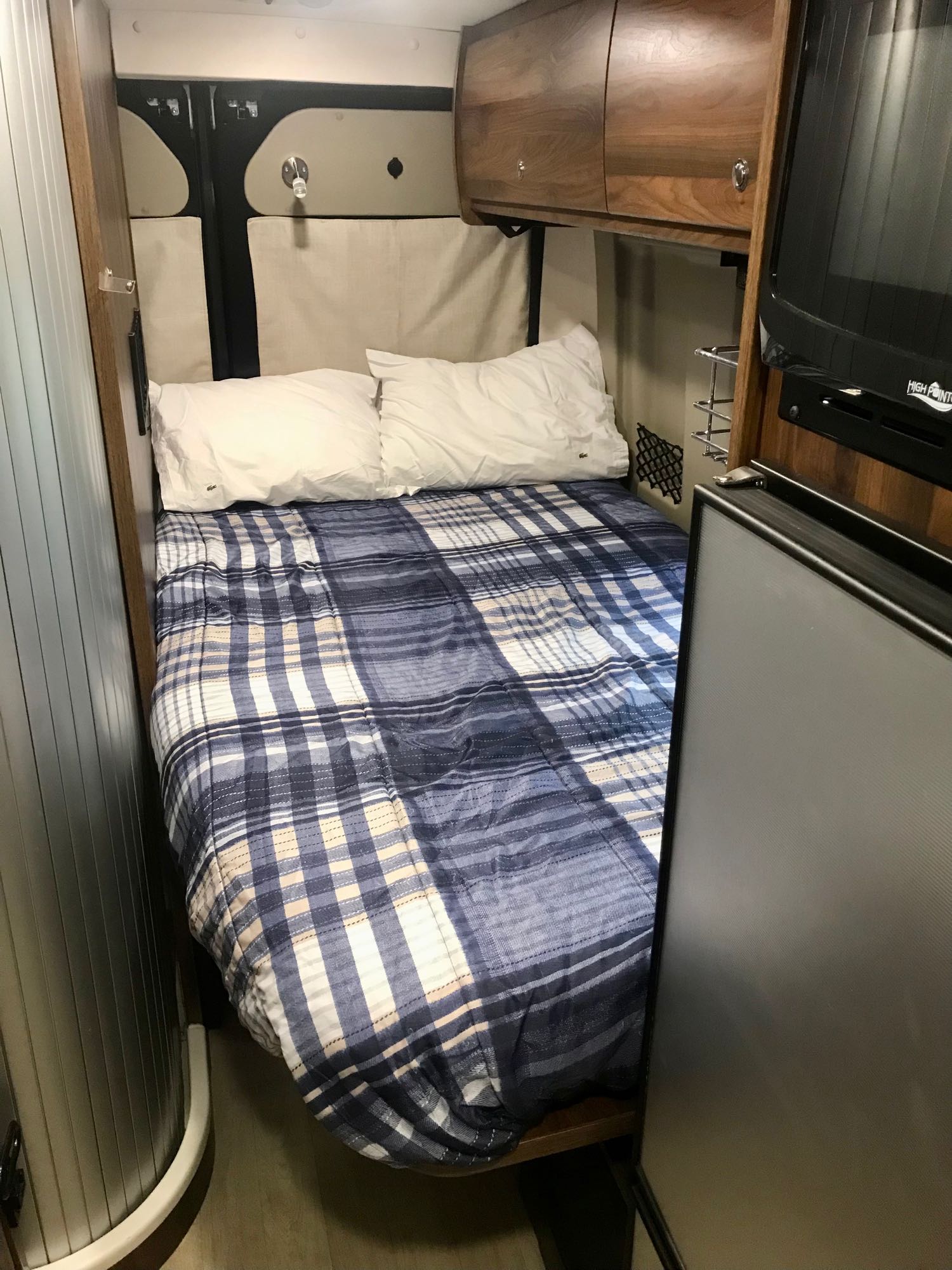 Cozy RV rental bedroom featuring a plaid bed, overhead cabinets, TV, and fridge. Ideal compact sleeping area.. winnebago Travato 59GL 2020