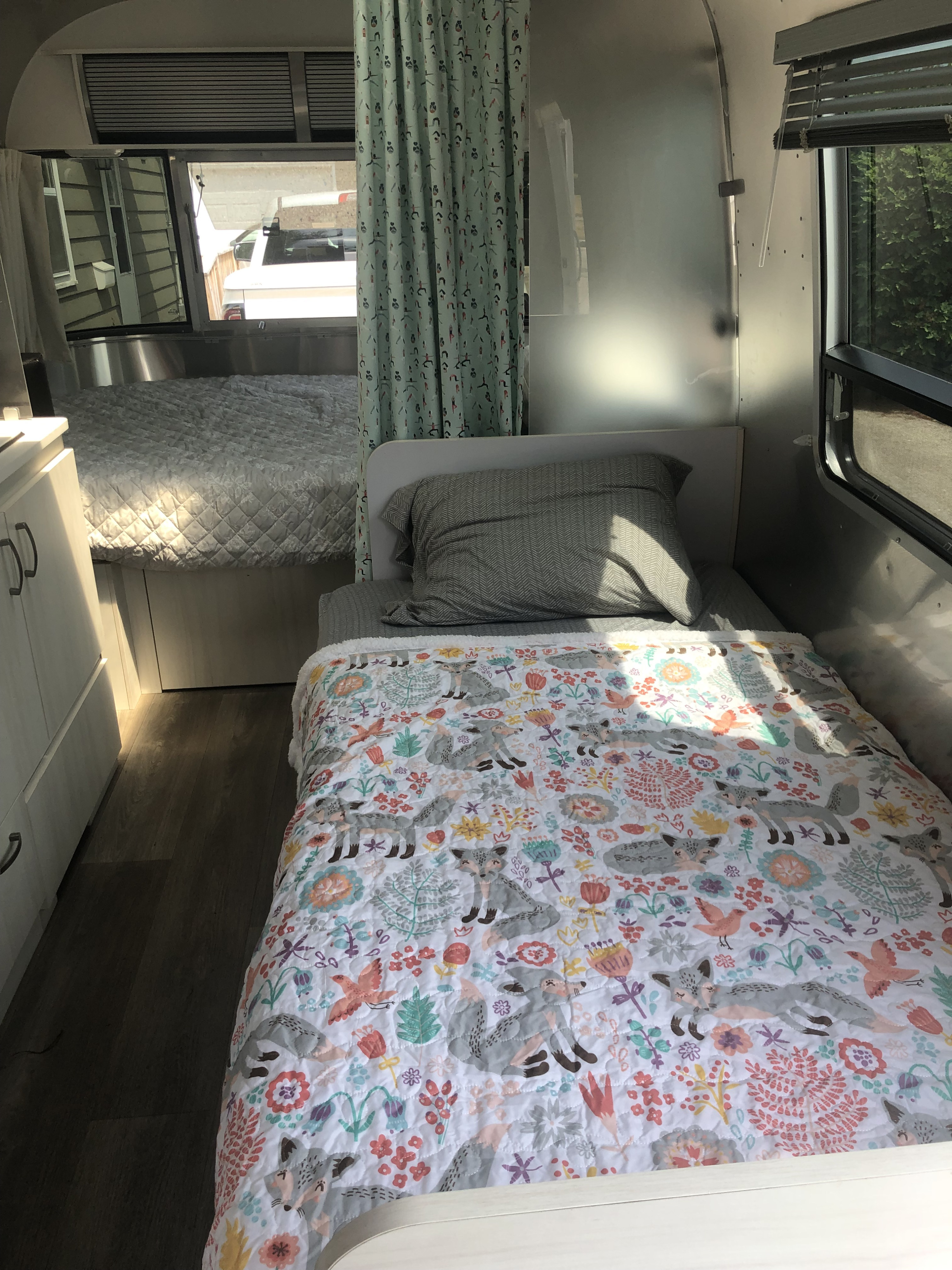Bright rv rental interior, featuring a twin bed with a whimsical fox quilt and a larger bed beyond. Shiny walls reflect light.. Airstream Sport 2019