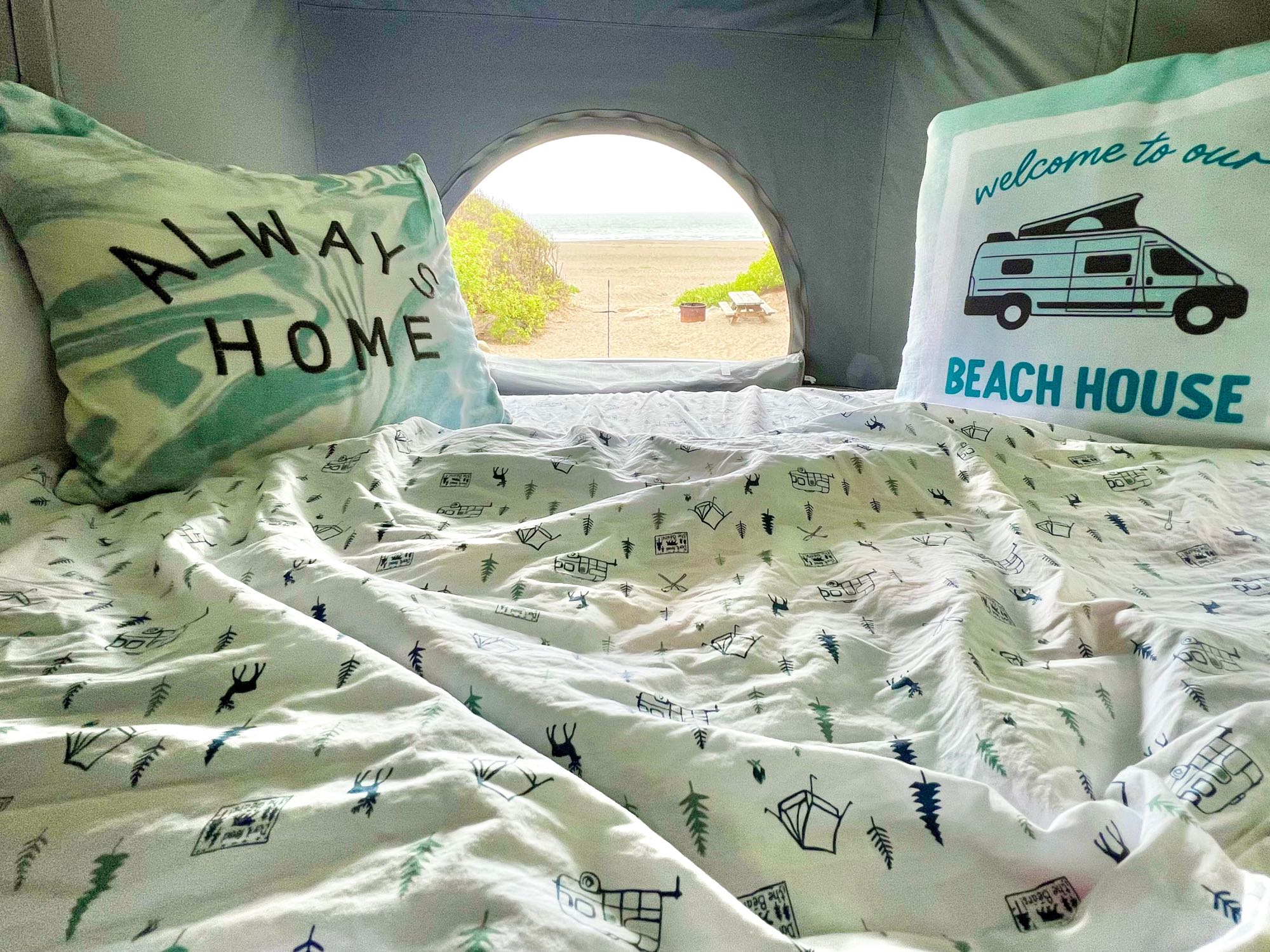 Cozy RV rental bed with pillows ('ALWAYS HOME', 'BEACH HOUSE') facing an arched window that overlooks a sandy beach.. Winnebago Solis BUT59P 2024