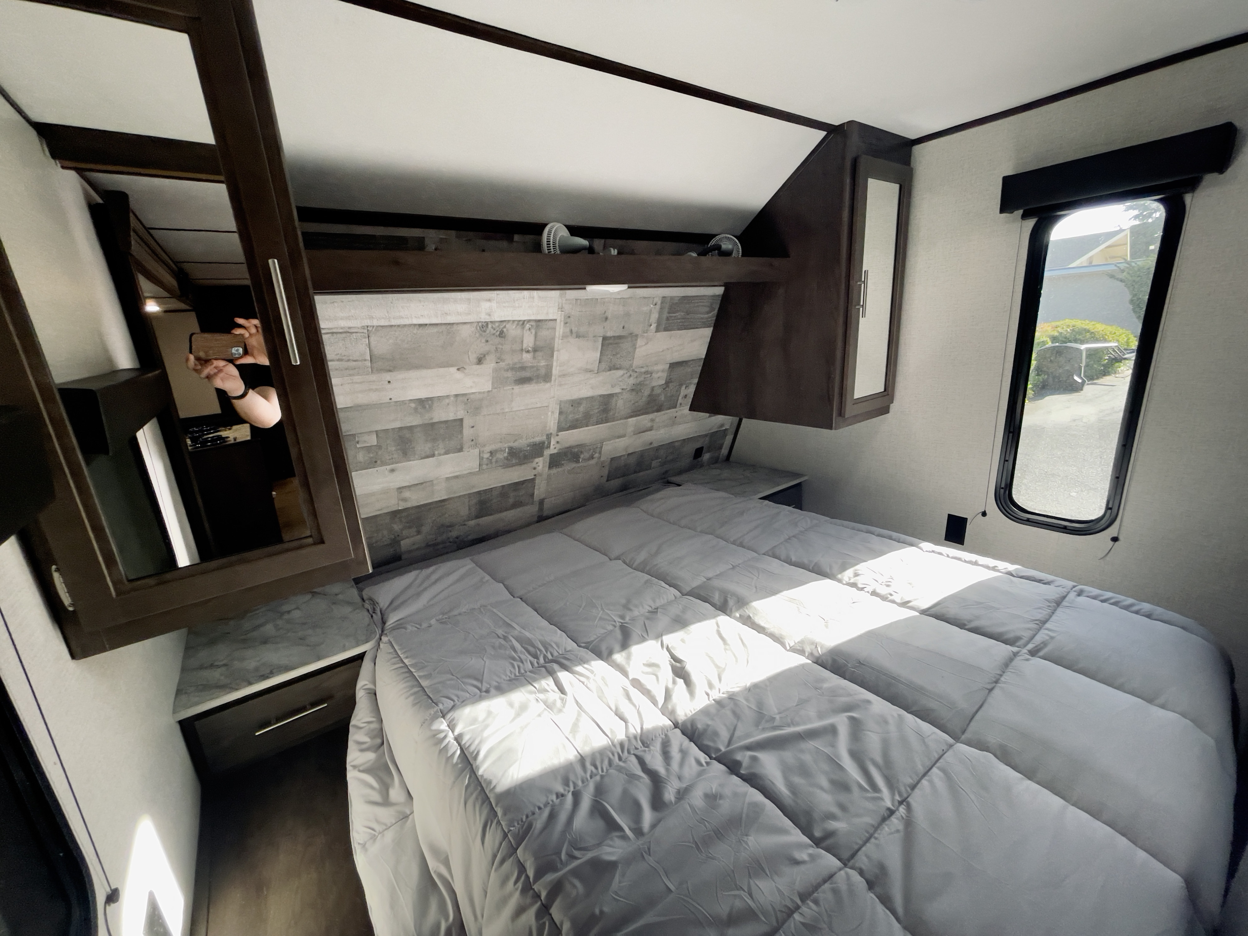Cozy RV rental bedroom with light gray bed, wood-paneled headboard, and built-in storage. Sunlight enters through window.. Dutchmen Coleman Lantern 2021