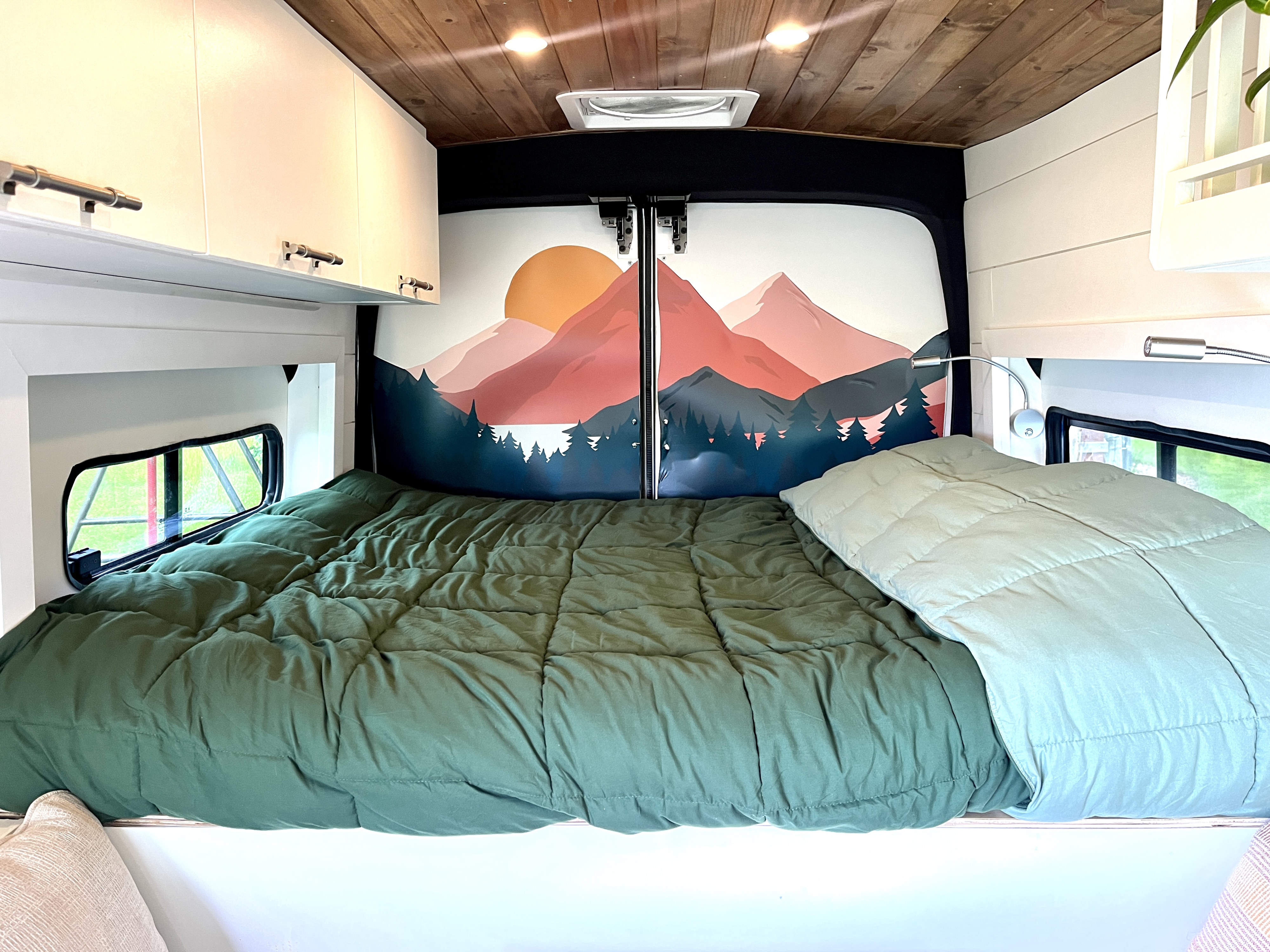 RV rental bedroom: a bed with green and light blue comforters, a vibrant mountain mural, white cabinets, and wooden ceiling.. Ford Transit 2017