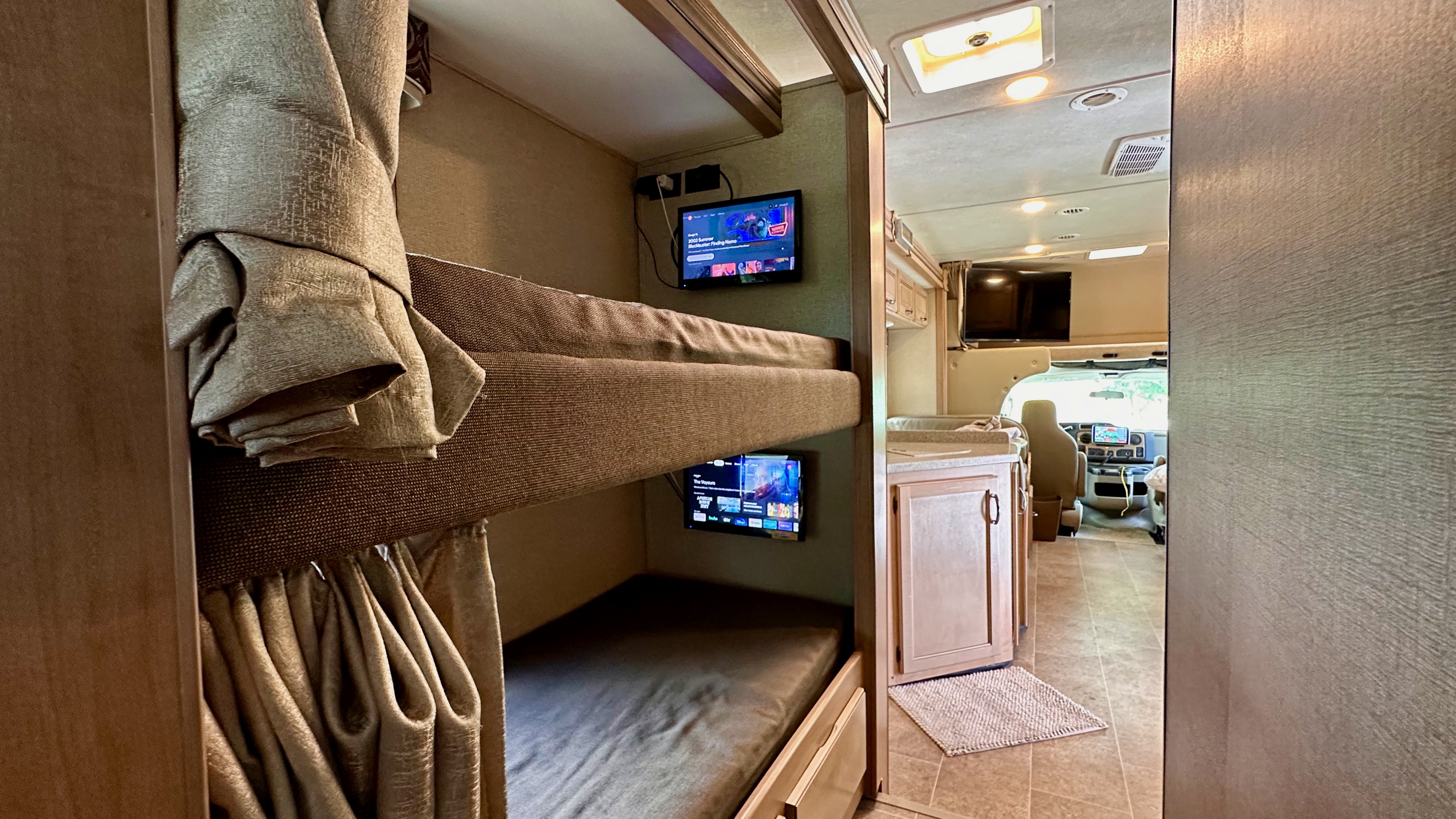 This RV rental interior features cozy bunk beds with personal TVs, leading to a bright, spacious living area toward the driver's cab.. Thor Chateau 2018