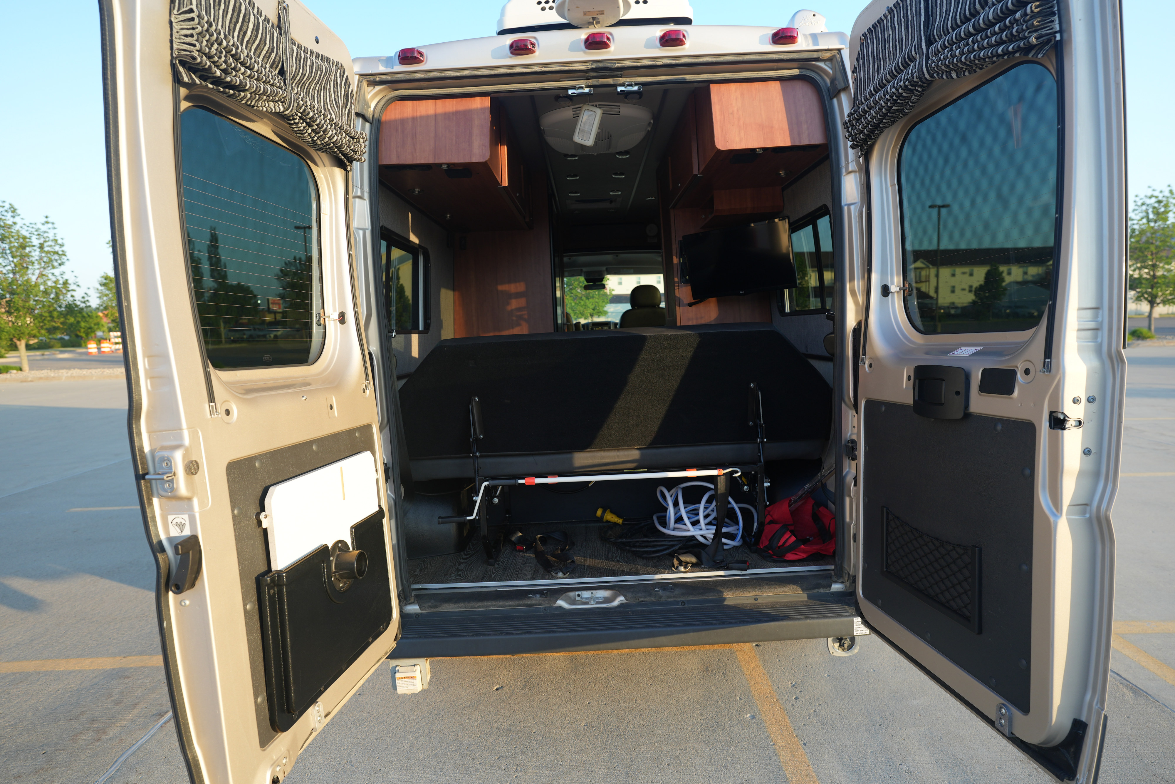 View into an RV rental from the open rear doors, showing the bed, overhead cabinets, TV, and stored items.. Roadtrek Roadtrek Class B 2018