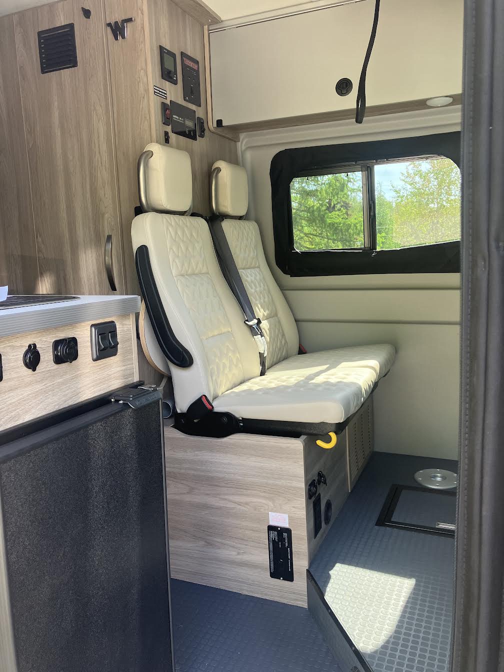 RV rental interior showcases two cream quilted seats converting to a bed, a fridge, control panels, and a window.. Winnebago Winnebago 2023