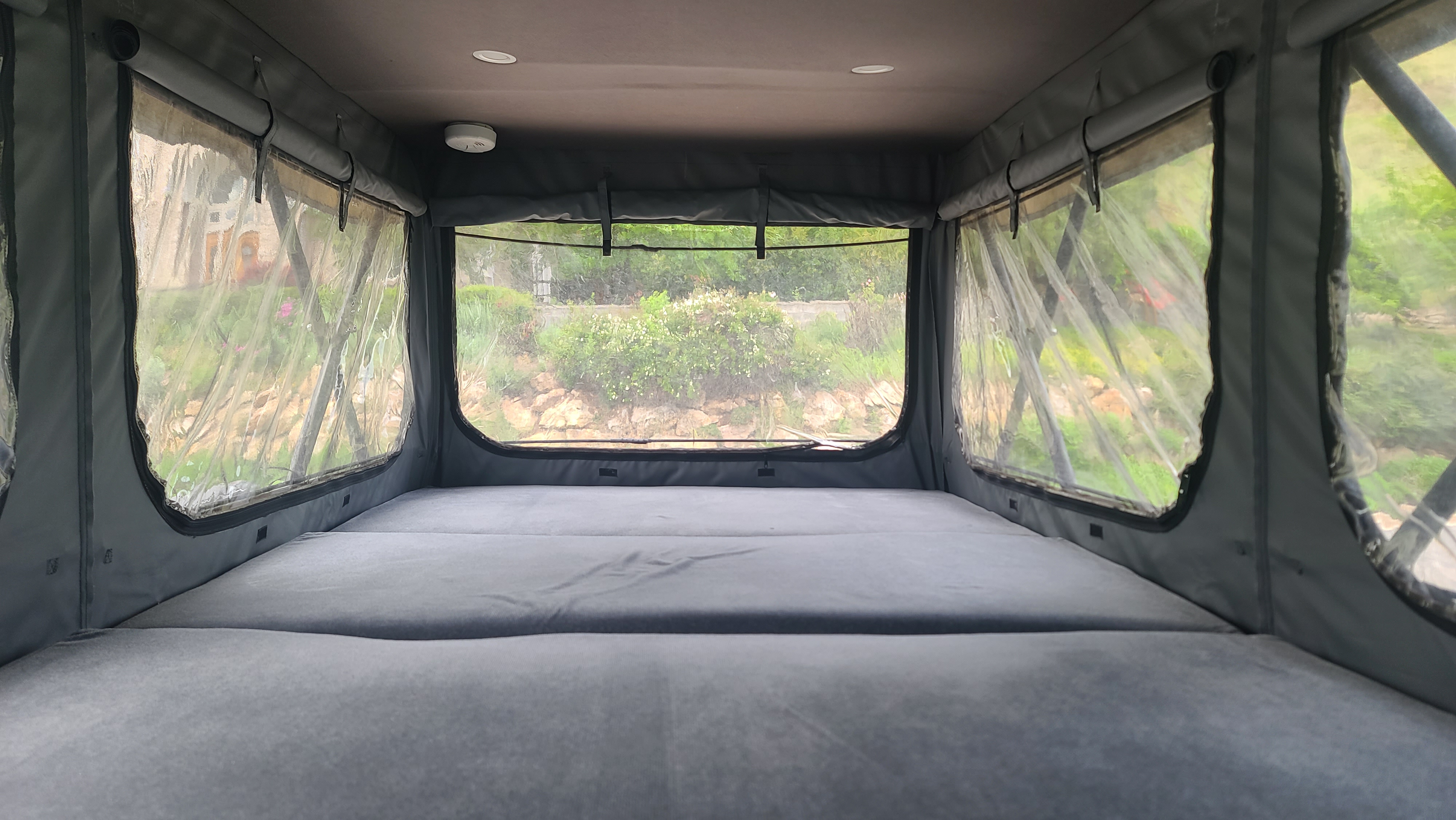 Spacious RV rental pop-top with a gray mattress and clear panoramic windows revealing lush green outdoor scenery.. ModVans ModVans Class B 2021