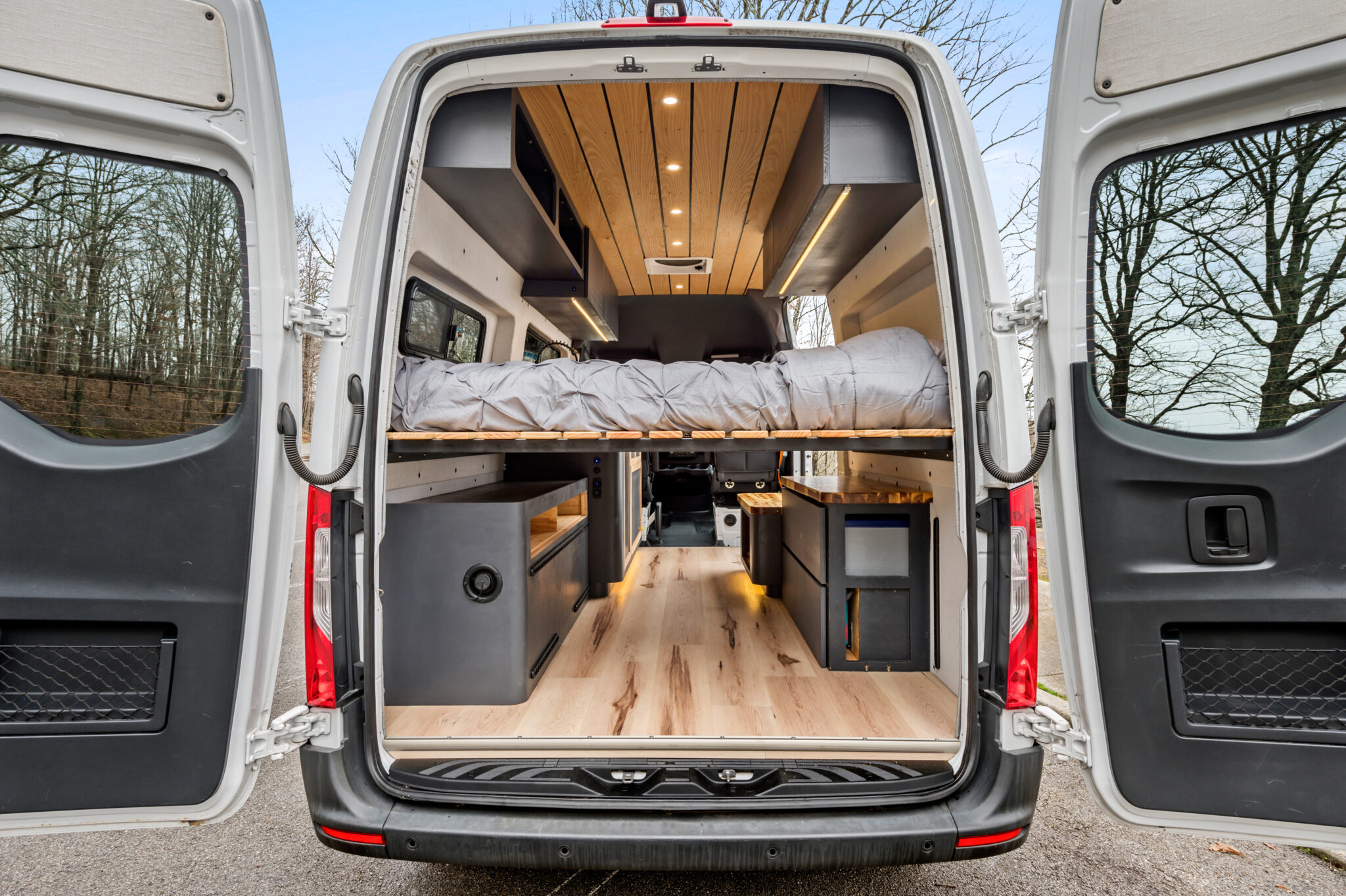 Rear interior of a custom camper van, revealing a raised bed, wood flooring, and modern cabinetry, ideal for rv rental adventures.. Mercedes-Benz Sprinter 2019