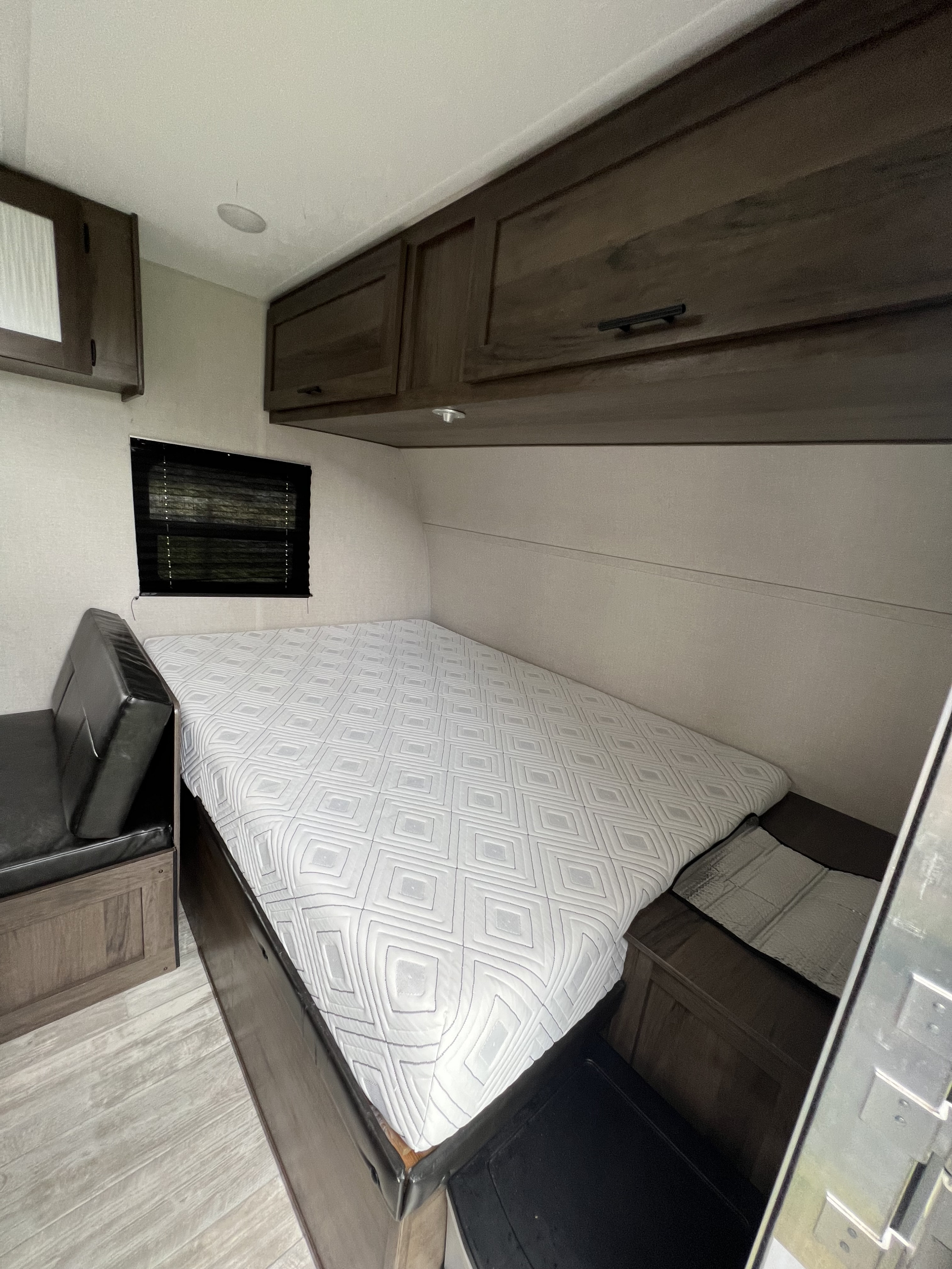 RV rental bedroom features a white patterned mattress on a bed base, dark wood overhead cabinets, and a window with blinds.. Avenger LT 2021