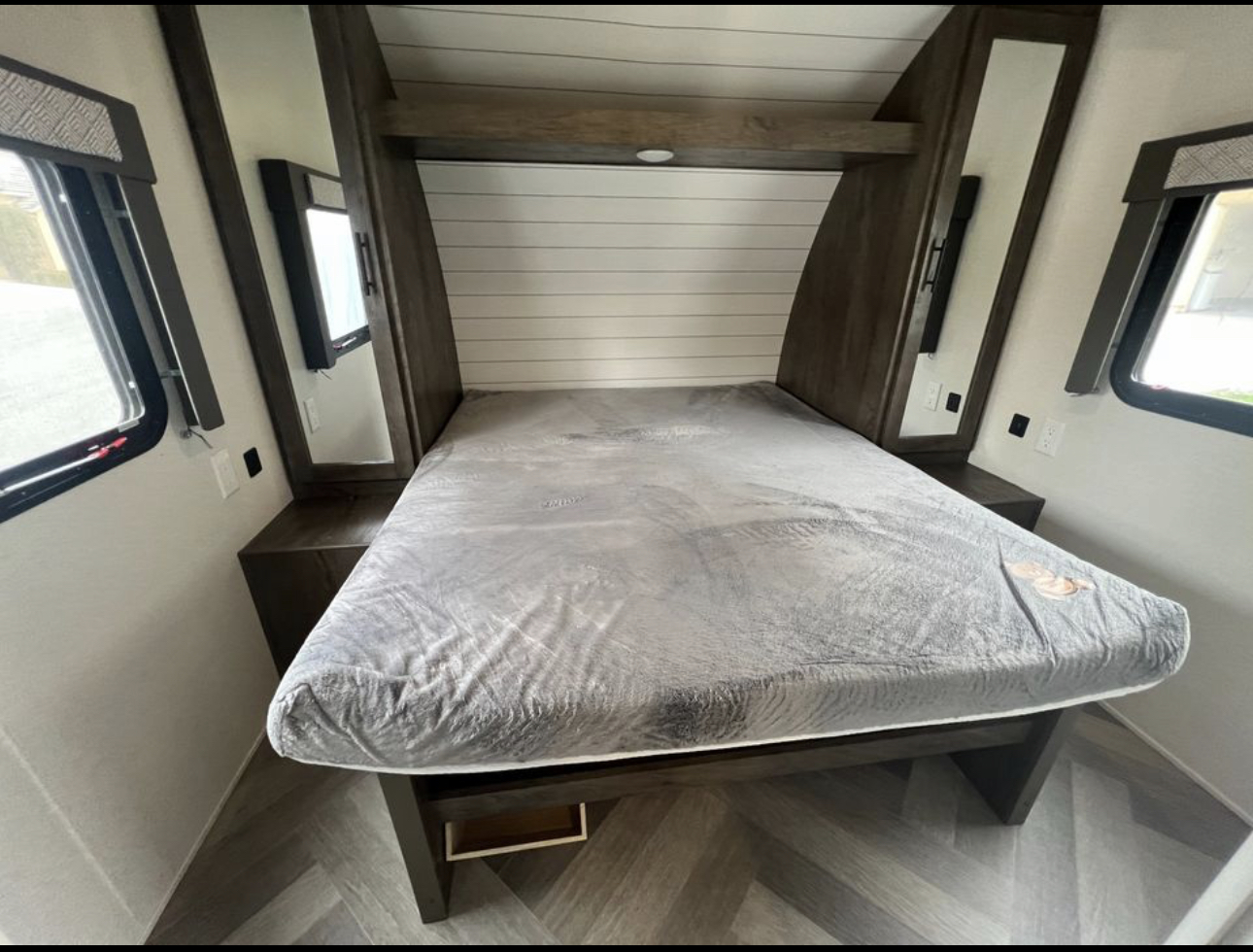 Step into this modern RV rental bedroom: comfy bed, integrated nightstands with mirrors, overhead storage, and windows.. Forest River Wildwood 2021