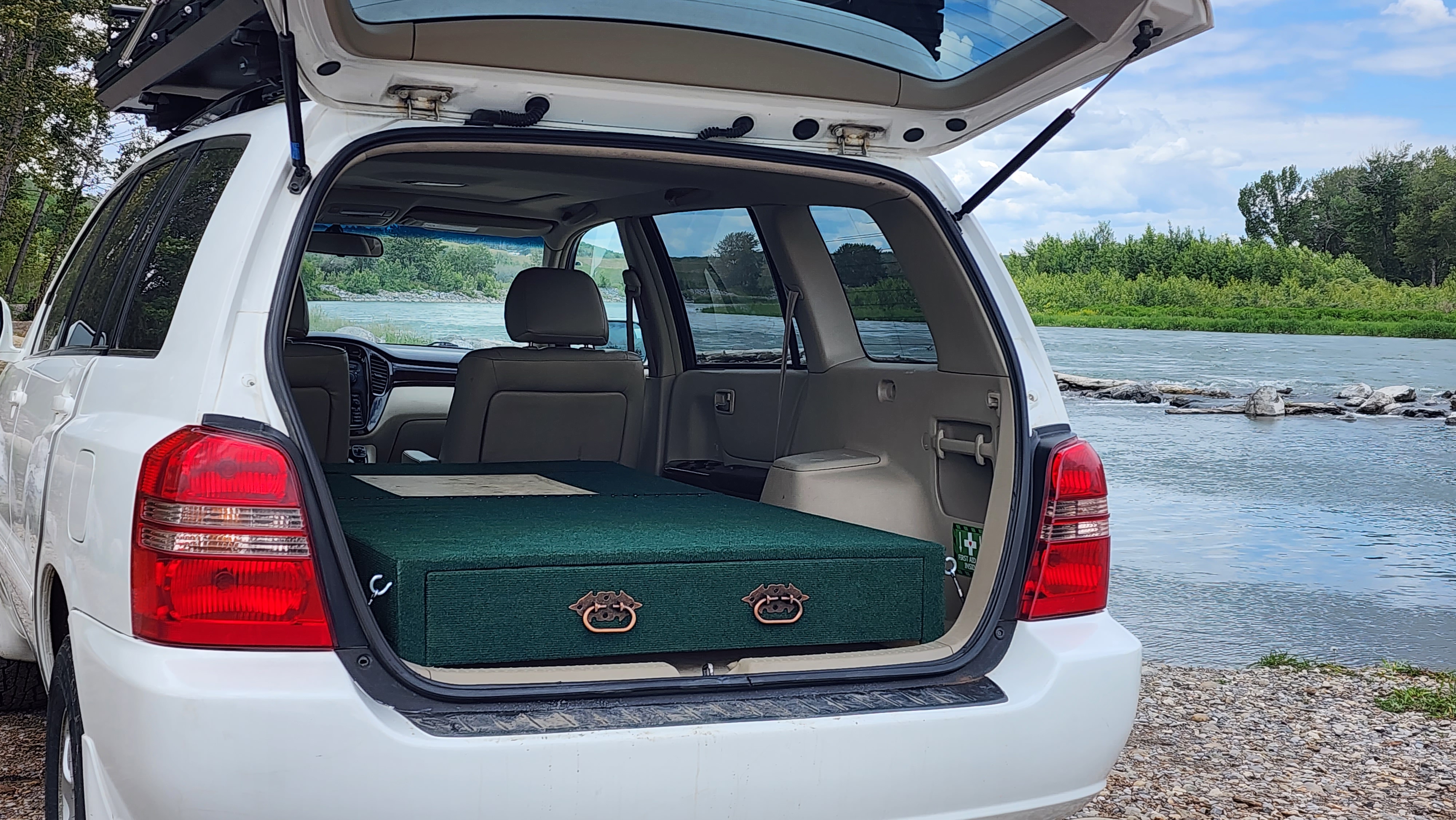 White SUV with custom green car camping sleeping platform and drawers by a river, offering an RV rental alternative.. Toyota Highlander 2005
