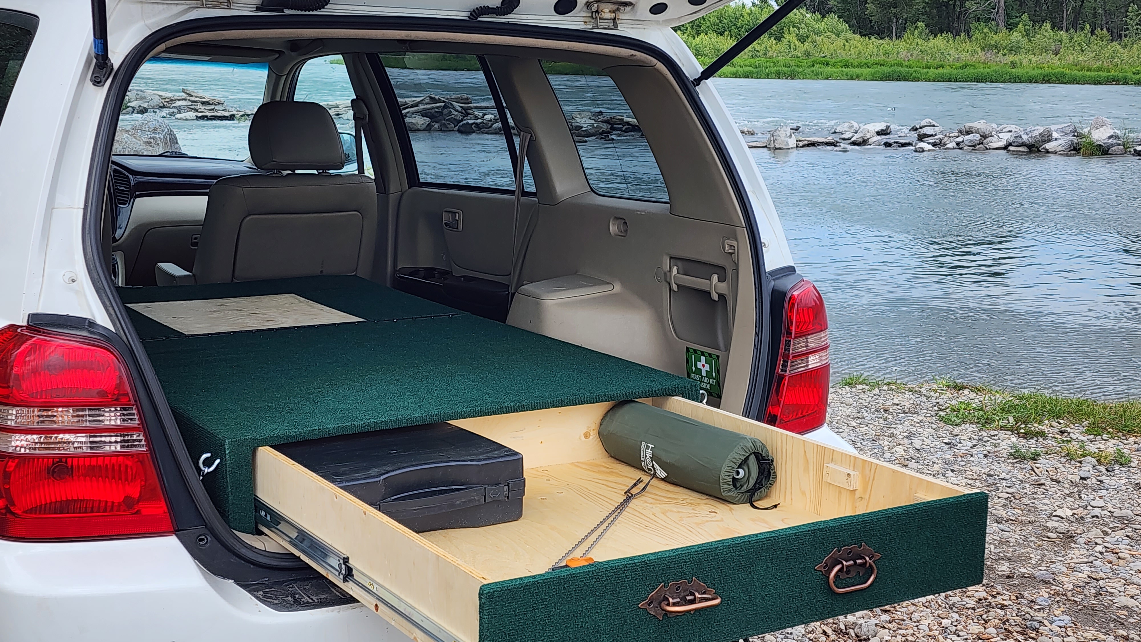 SUV car camping setup with a built-in bed platform and drawer by a river. An RV rental alternative for outdoor adventures.. Toyota Highlander 2005