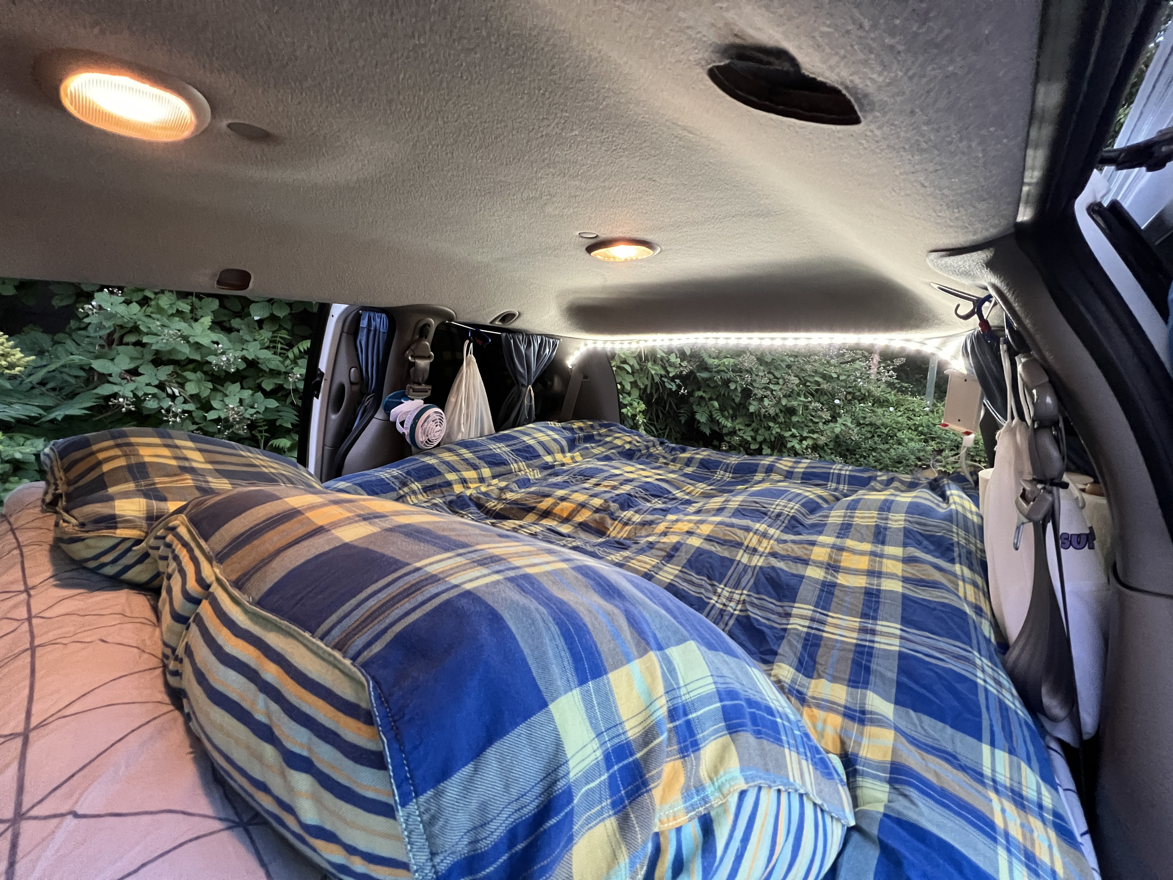 An RV rental's cozy sleeping area, featuring a blue plaid bed, warm interior lights, and lush greenery outside.. Dodge Caravan 2001