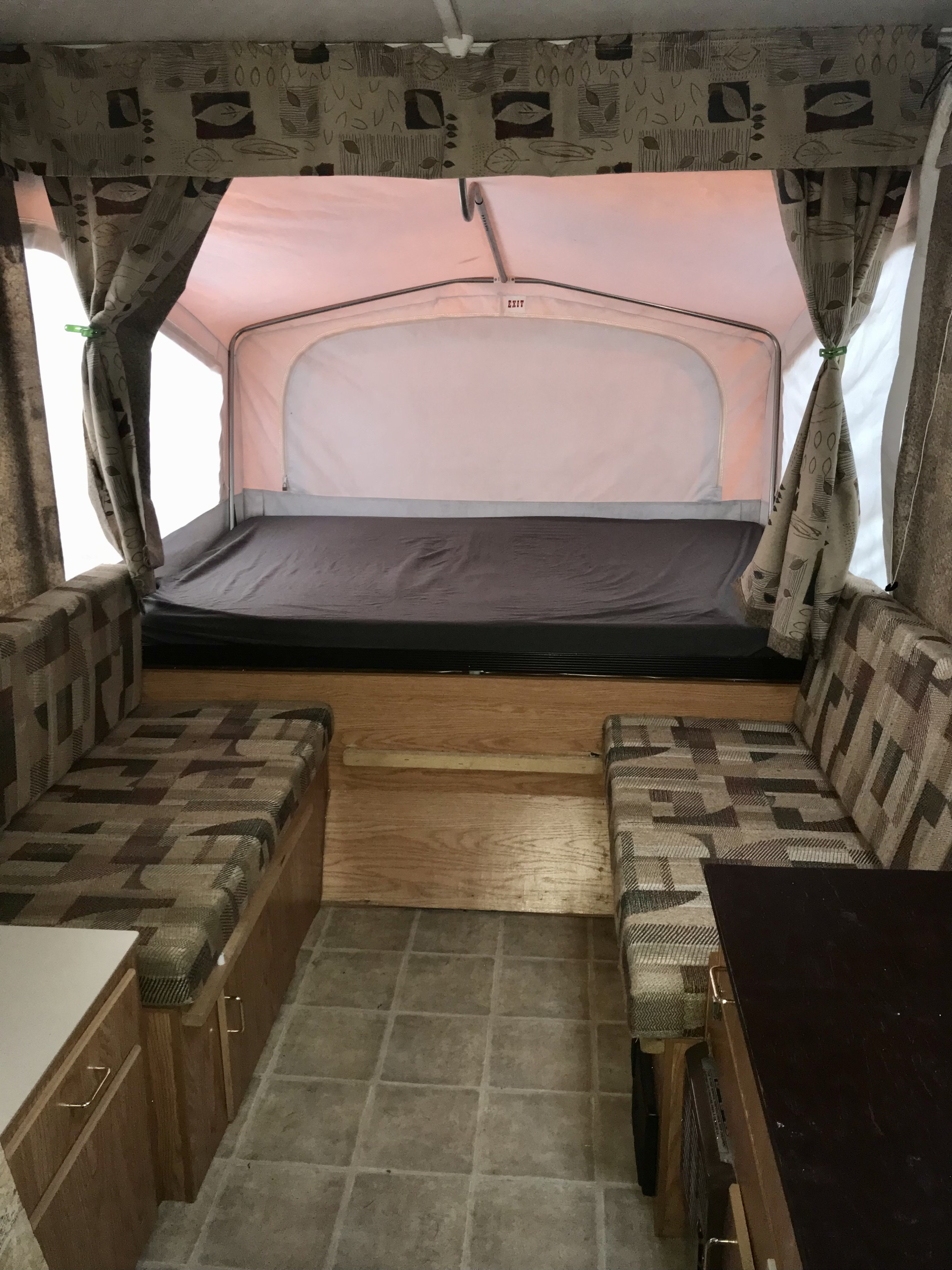 Inside this RV rental pop-up camper: a cozy bed, patterned seating benches, and a light-colored ceiling with curtains.. Jayco Qwest 2006