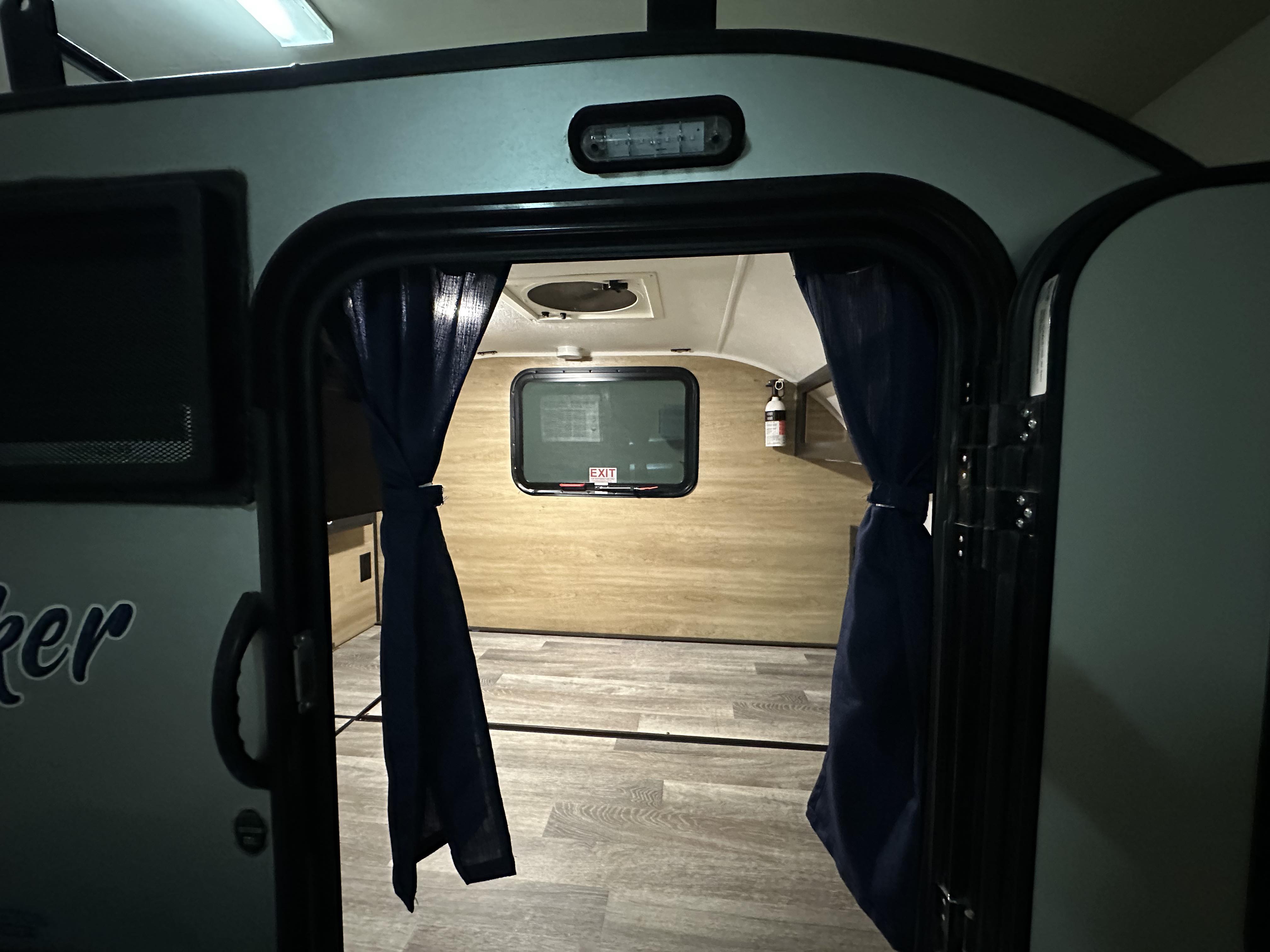 View inside an RV rental: light wood paneling, window with exit sign, fire extinguisher, and tied-back blue curtains.. Braxton Creek Bushwhacker 2021