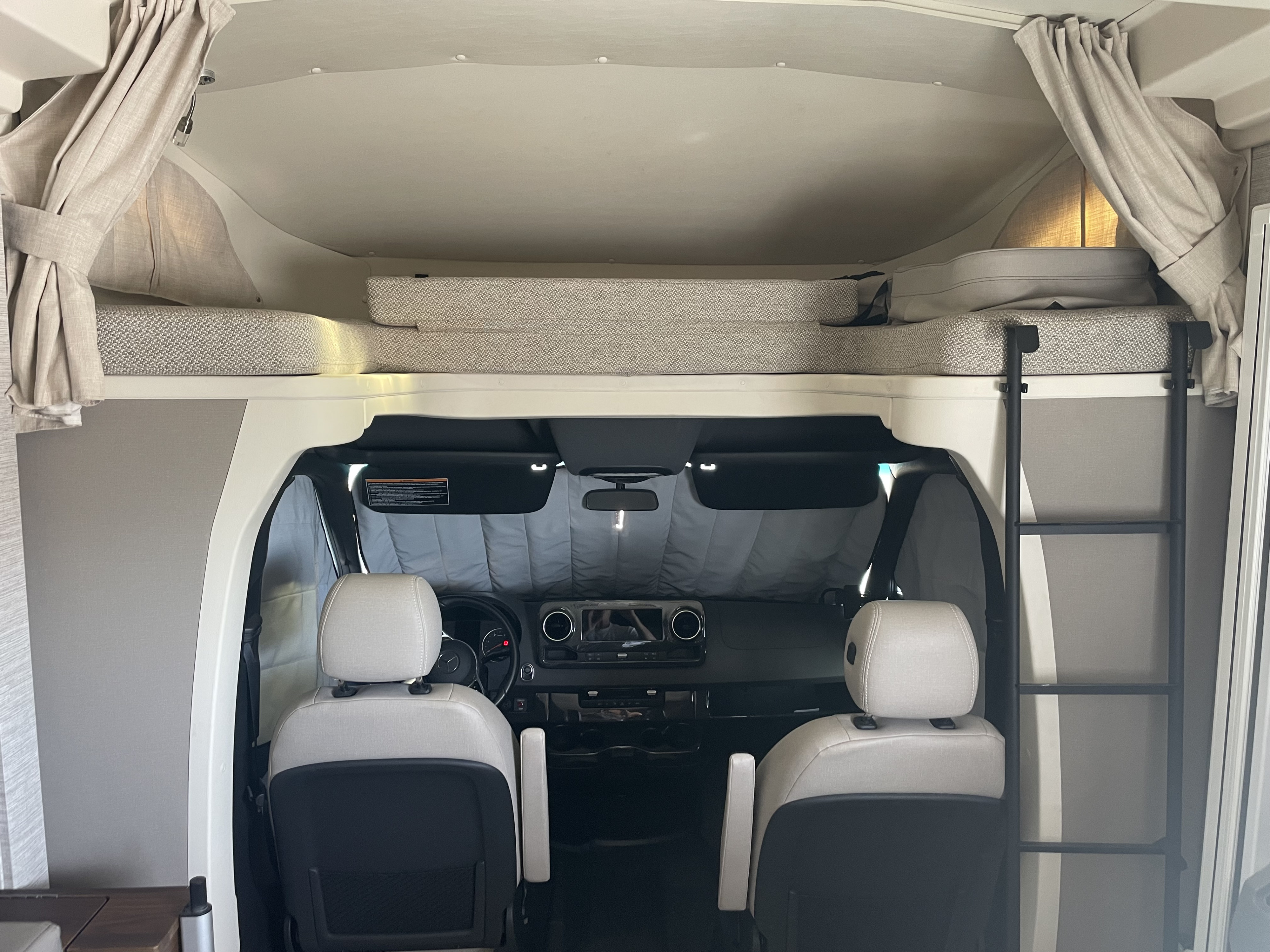 RV rental interior: over-cab bunk bed with ladder, and the front cockpit with a sunshade covering the windshield.. Winnebago Navion 2022