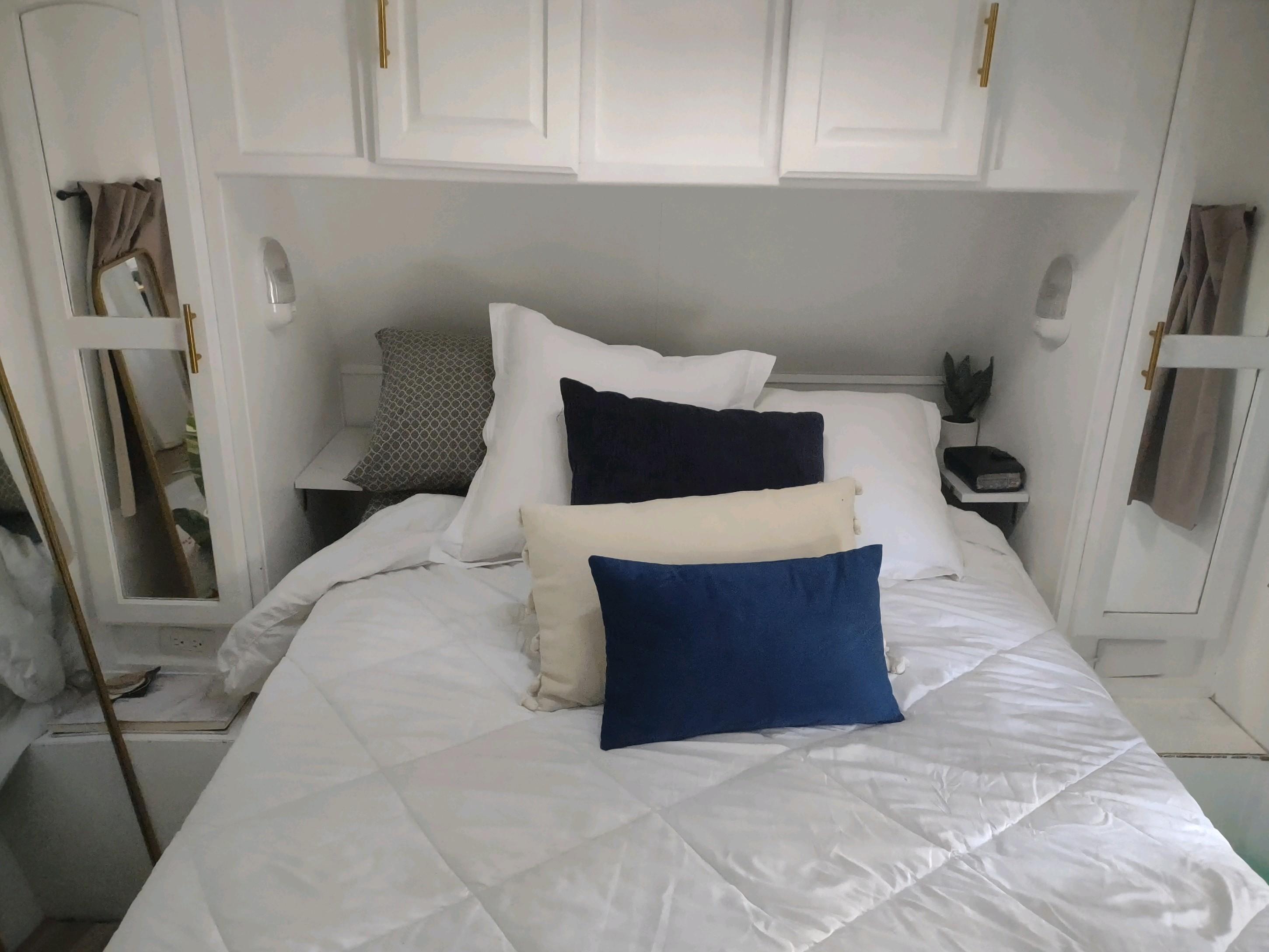 Cozy RV rental bedroom with white bedding, blue and cream pillows, and overhead cabinets.. Forest River Surveyor Sport 2012