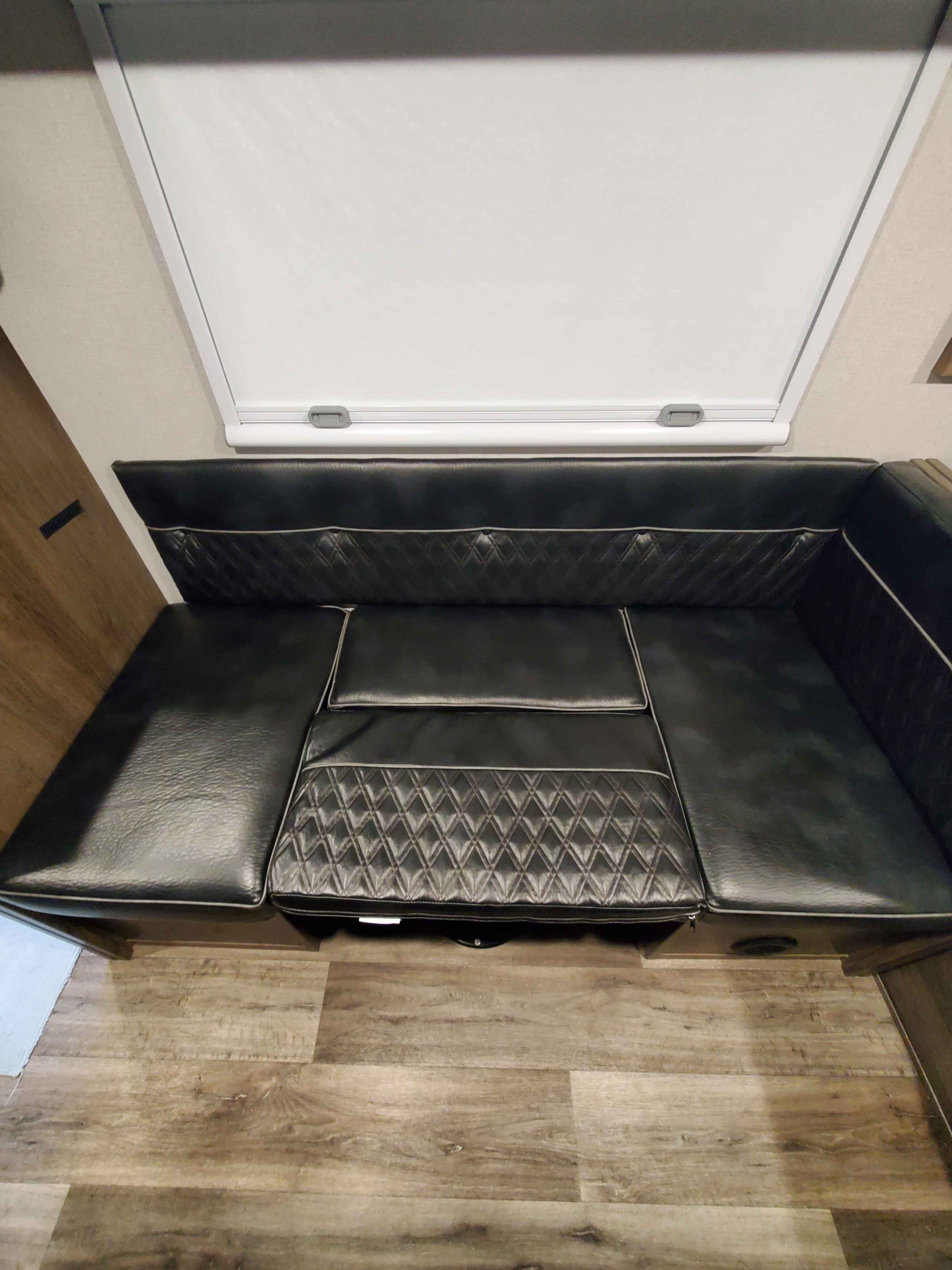 An L-shaped black faux leather RV dinette bench with a white window blind and wood-look flooring, great for any rv rental.. Imperial Outdoors XploreRV 2023
