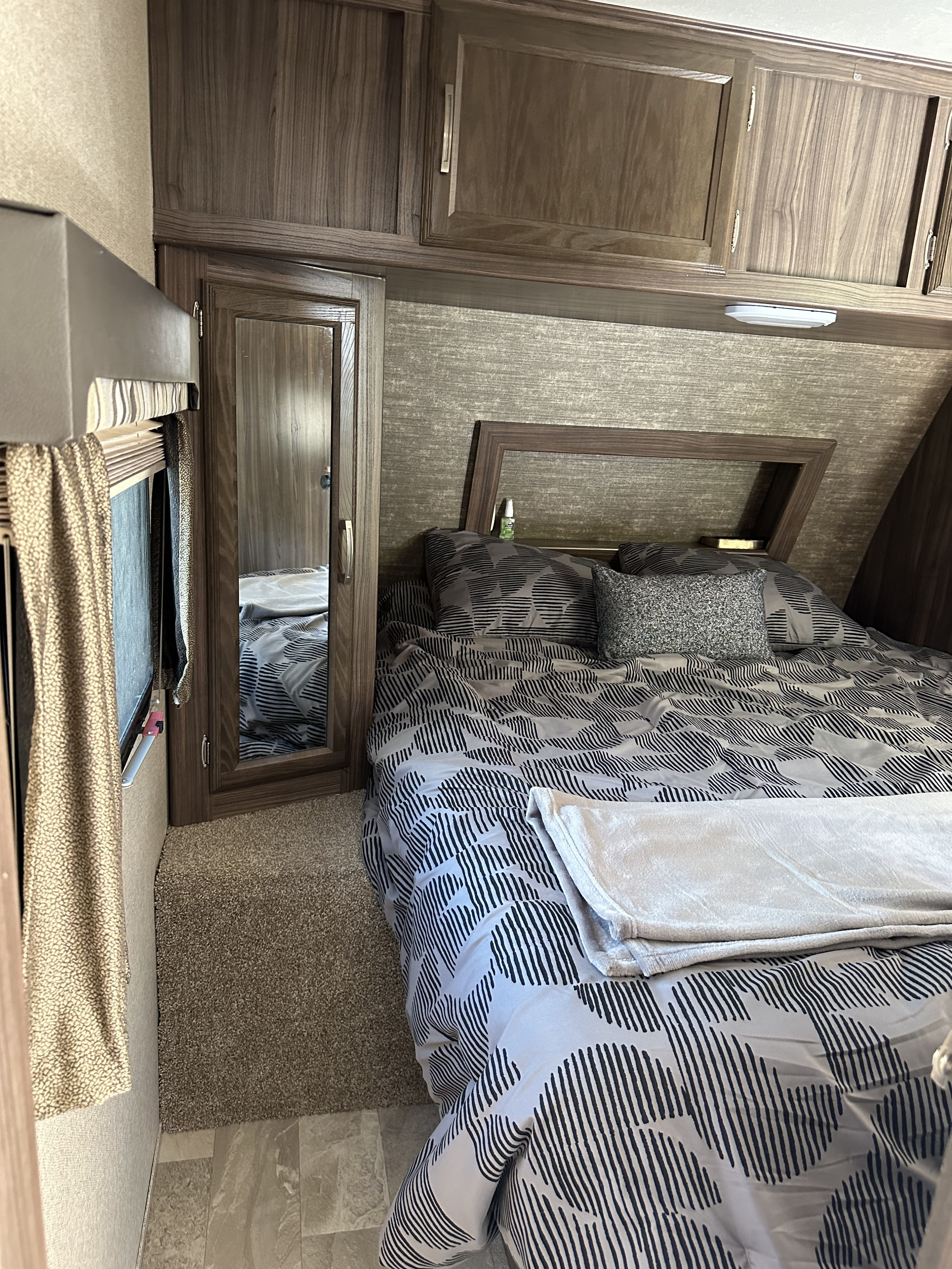 Cozy RV rental bedroom with patterned bedding, overhead wood cabinets, and a mirrored wardrobe. Provides comfortable sleeping space.. Coachmen Apex 2019