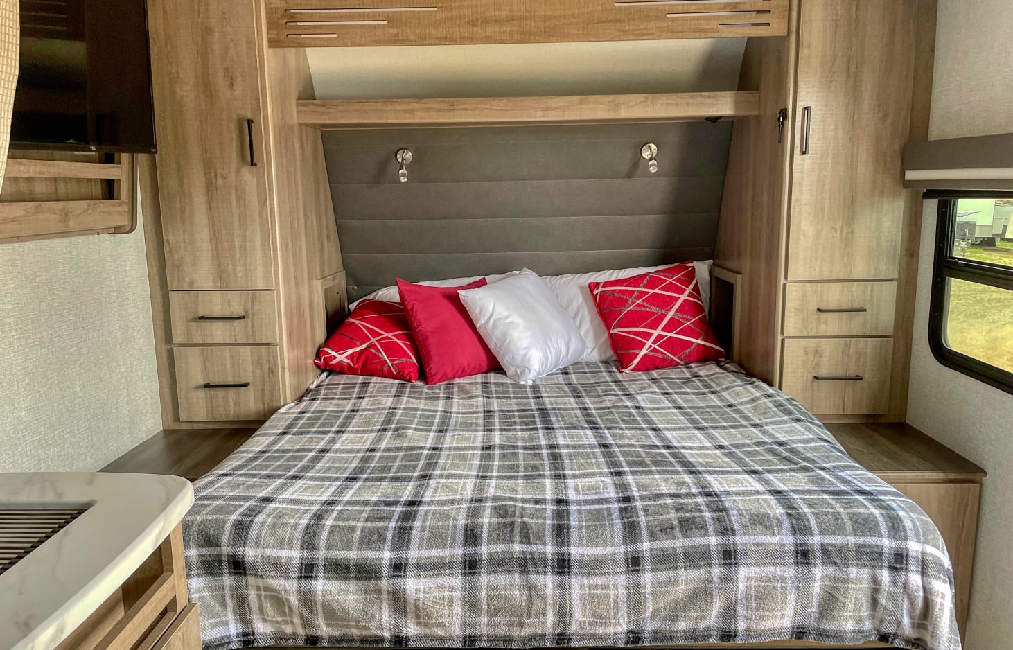 Cozy RV rental bedroom with a plaid blanket, red pillows, and wooden built-in storage. Features a TV and window.. Grand Design Other 2023