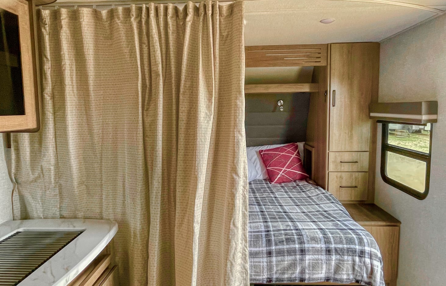Cozy RV rental bedroom with plaid bedding, a red pillow, and privacy curtain, ideal for travel trailer adventures.. Grand Design Other 2023