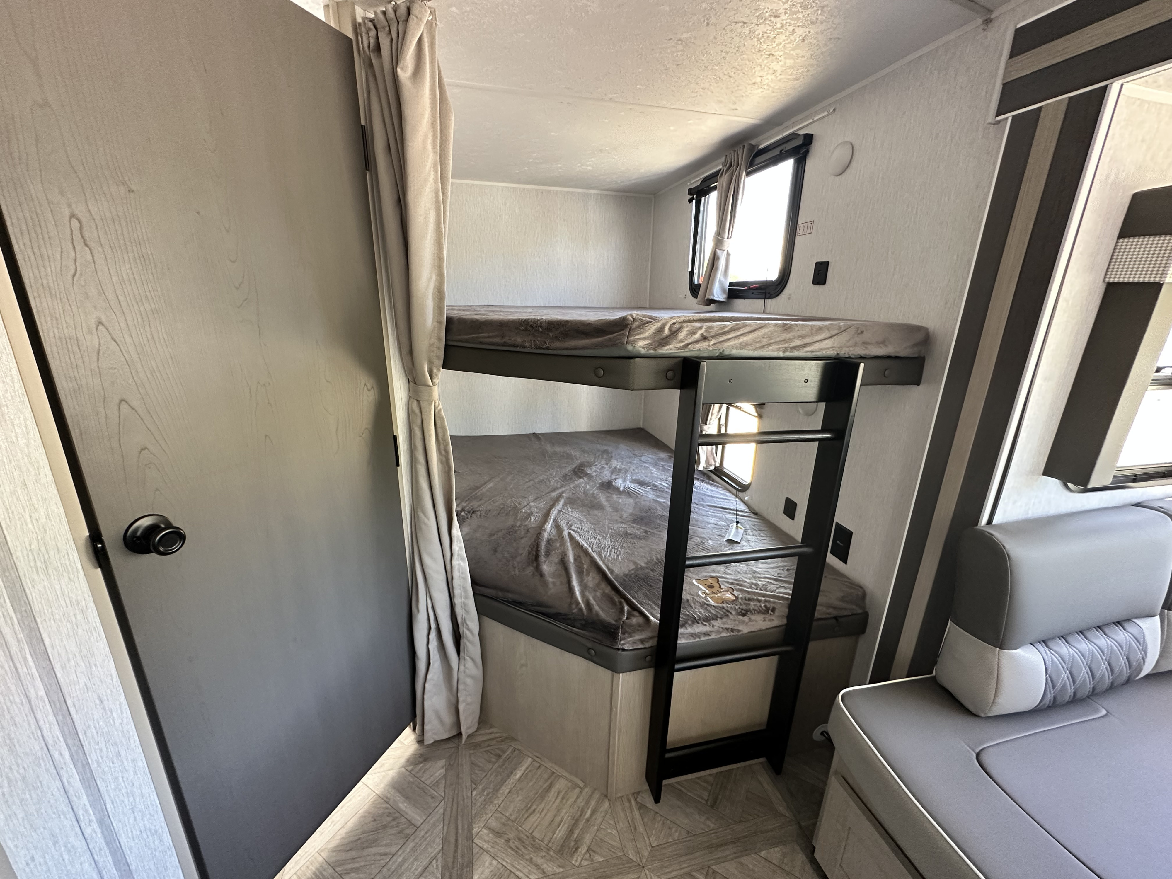 RV rental interior: bunk beds with grey bedding, ladder, window with curtain, and door. Textured walls, patterned floor, and seating.. Forest River Salem Cruise Lite 2023