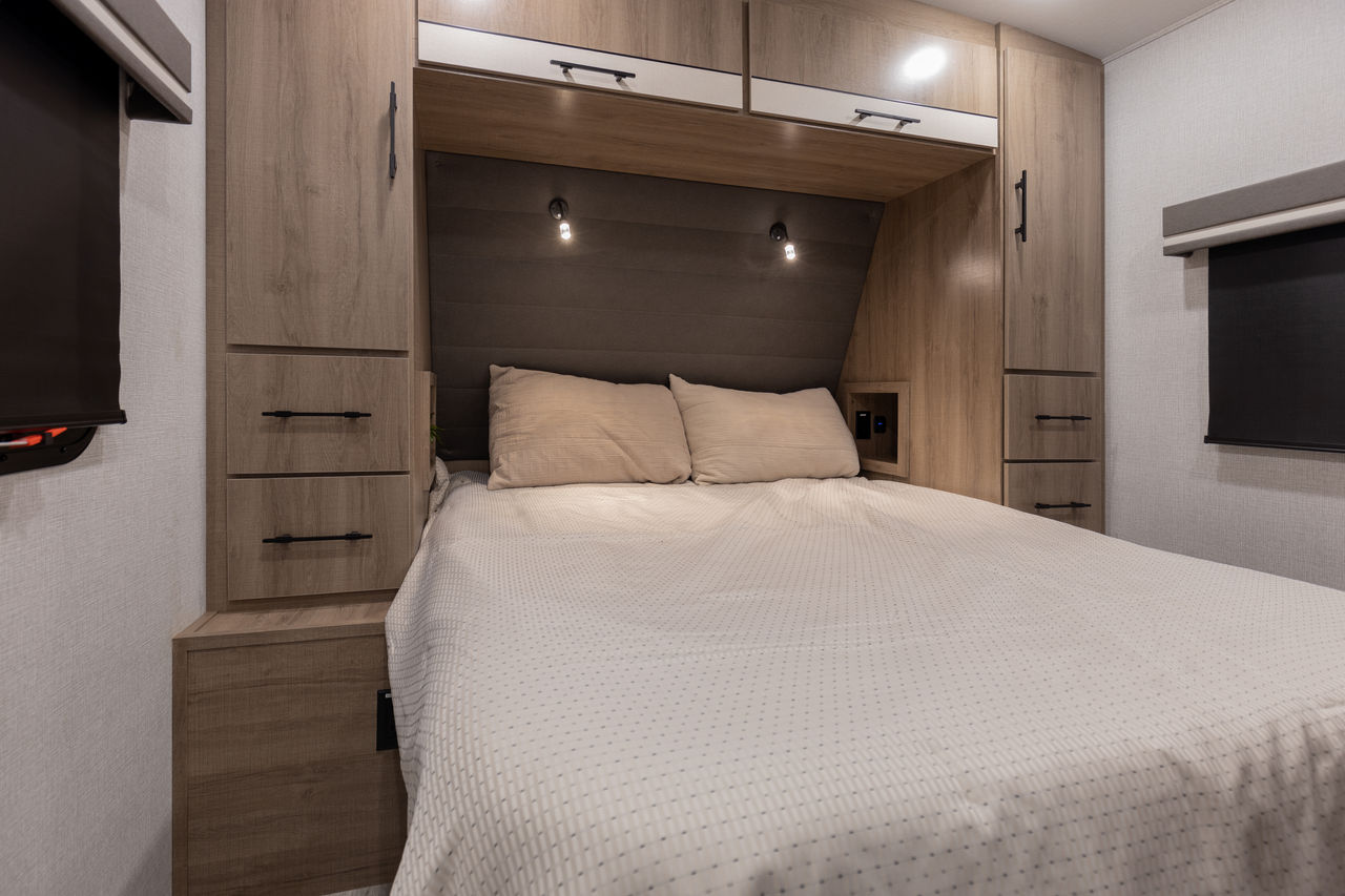 Compact RV rental bedroom featuring a bed, upholstered headboard, reading lights, and wood storage.. Grand Design Imagine 2023