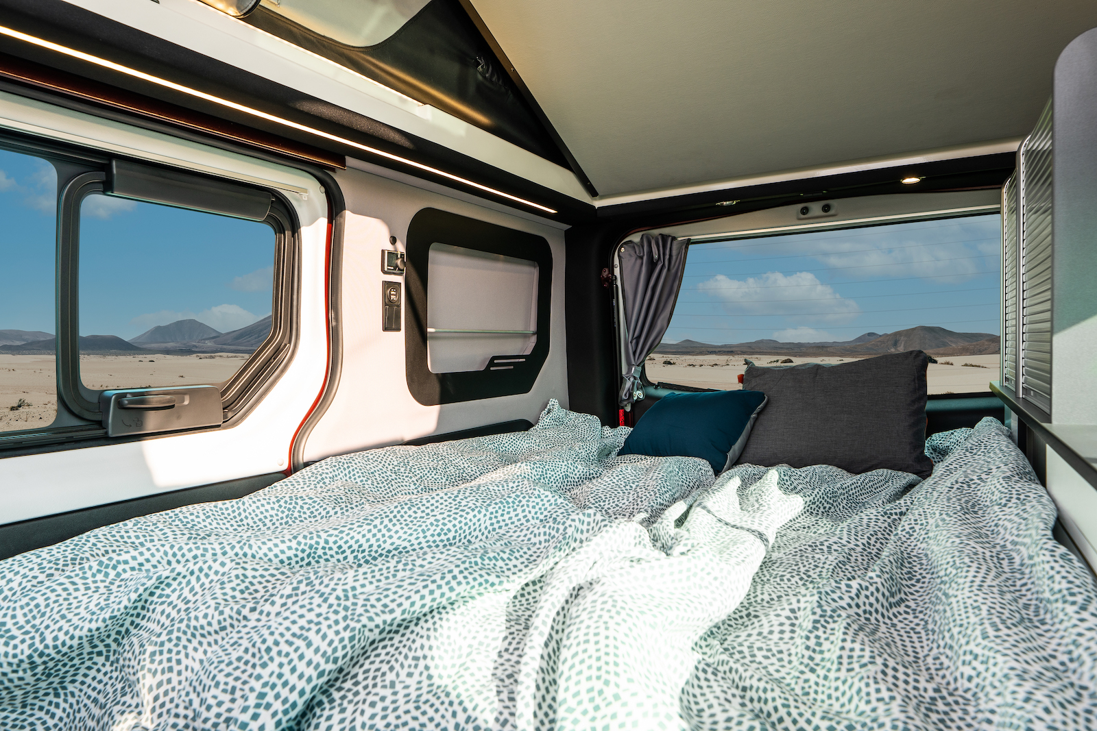 This RV rental features a cozy bed with patterned sheets, offering panoramic desert mountain views from its large windows.. HANROAD TREK 4 2023