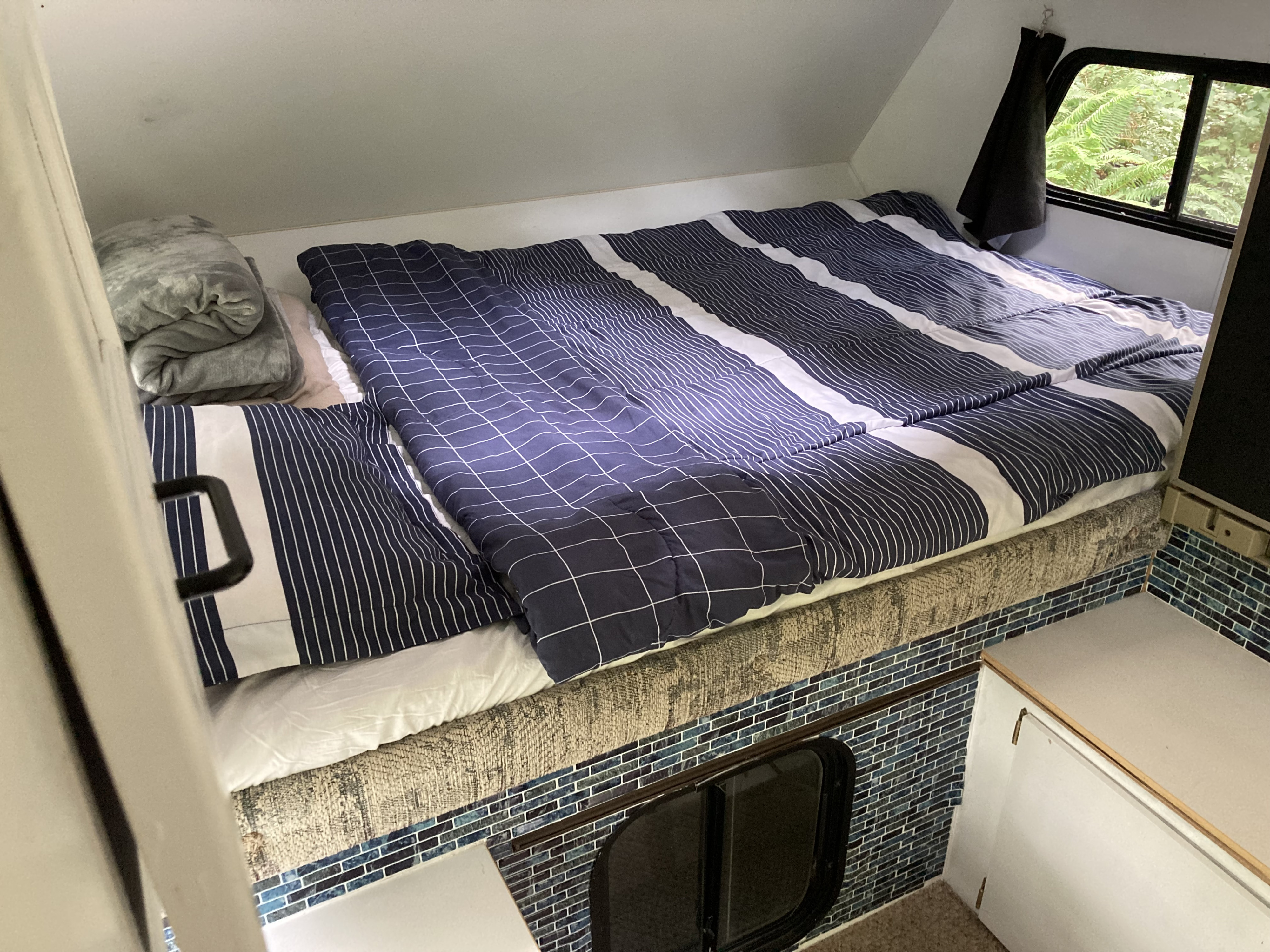 Cozy RV rental bed with blue striped bedding, a small window, and mosaic tile base. Ideal compact sleeping area.. Gmc Other 2003