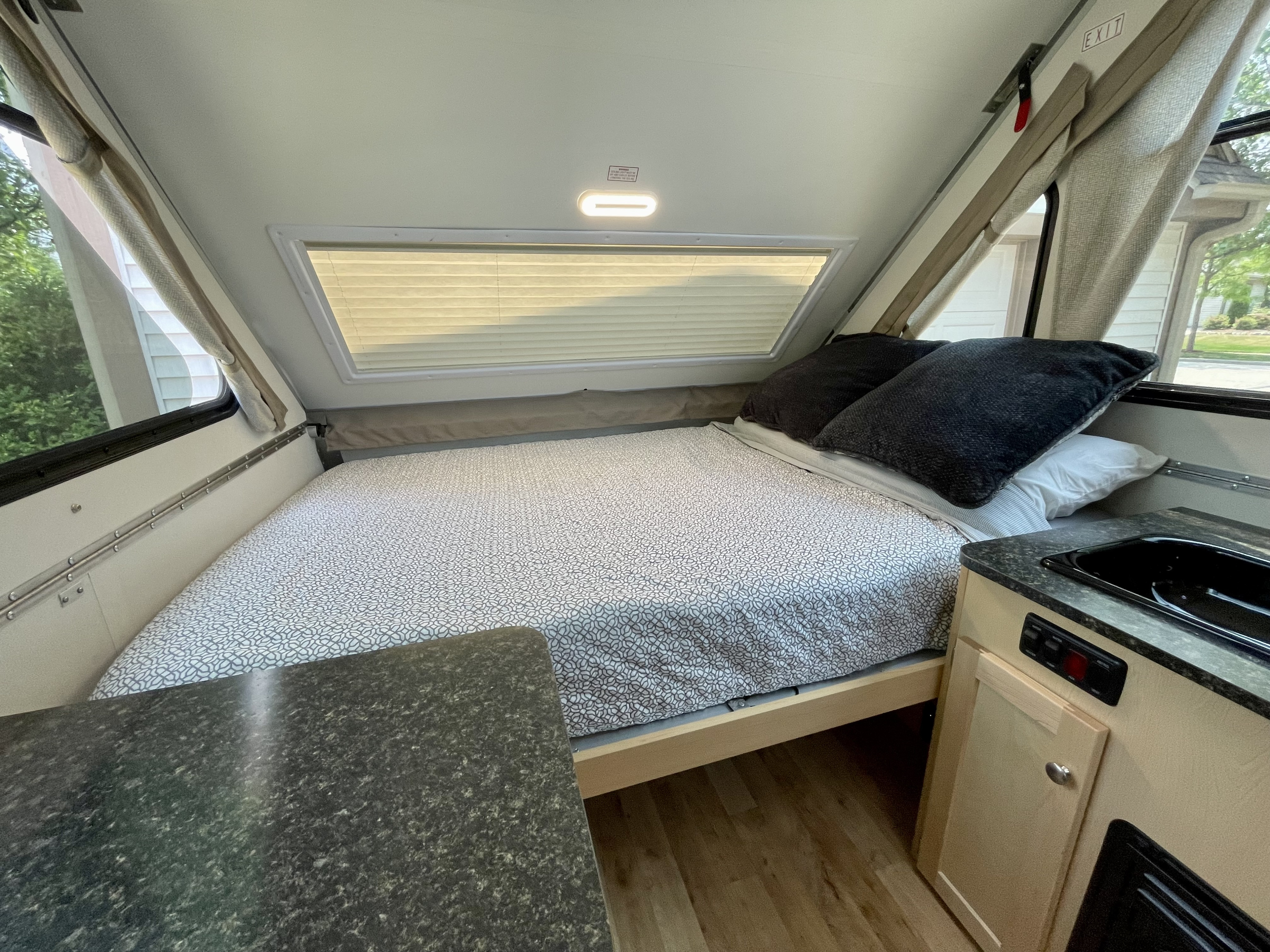 Inside a cozy RV rental: a comfortable bed with patterned comforter, window blinds, and a compact kitchen counter.. A Liner A Liner Popup Trailer 2023