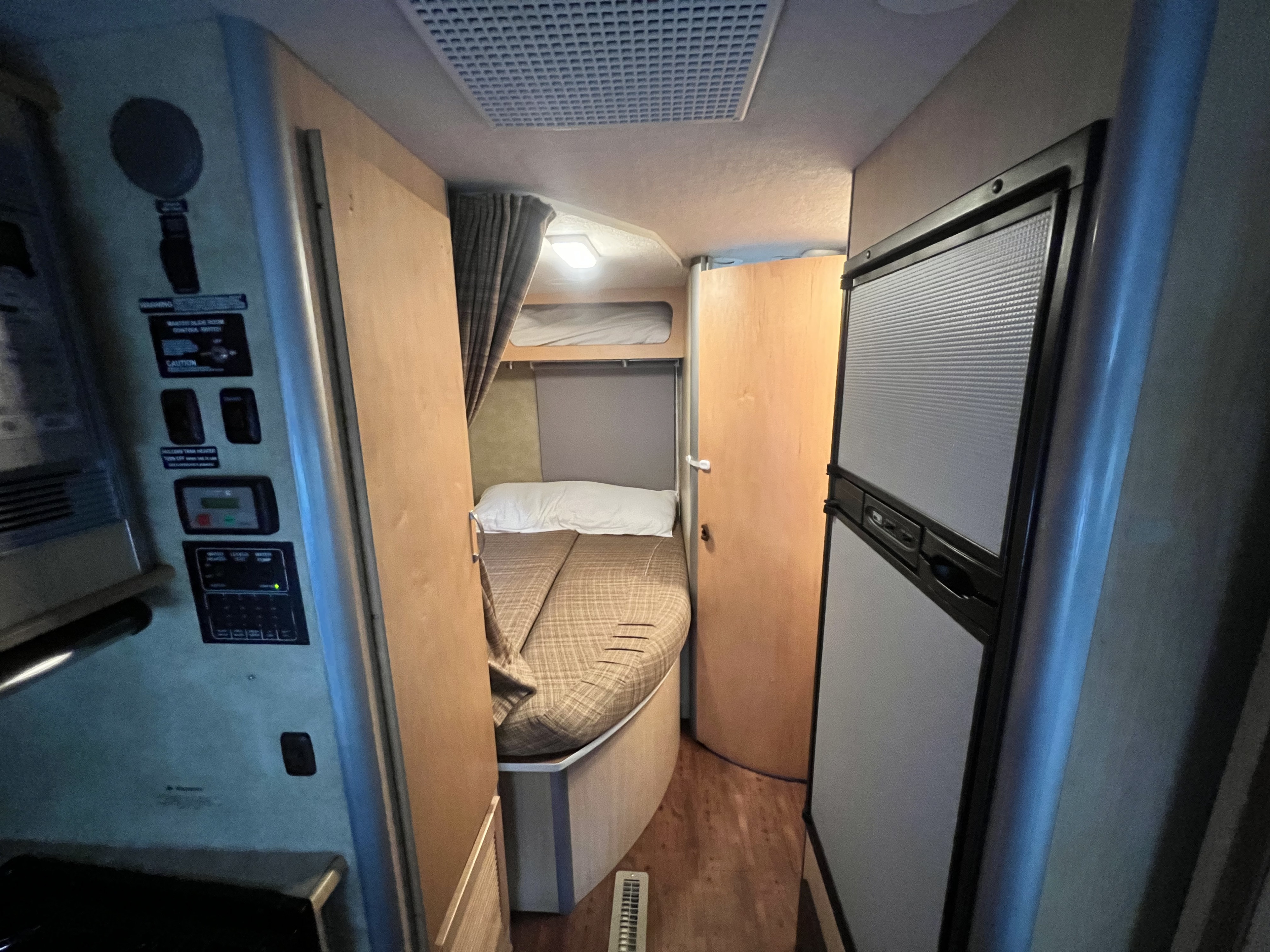 Inside an RV rental: a narrow hallway with a control panel and a full bed, plus an upper bunk and refrigerator.. Winnebago View 2008