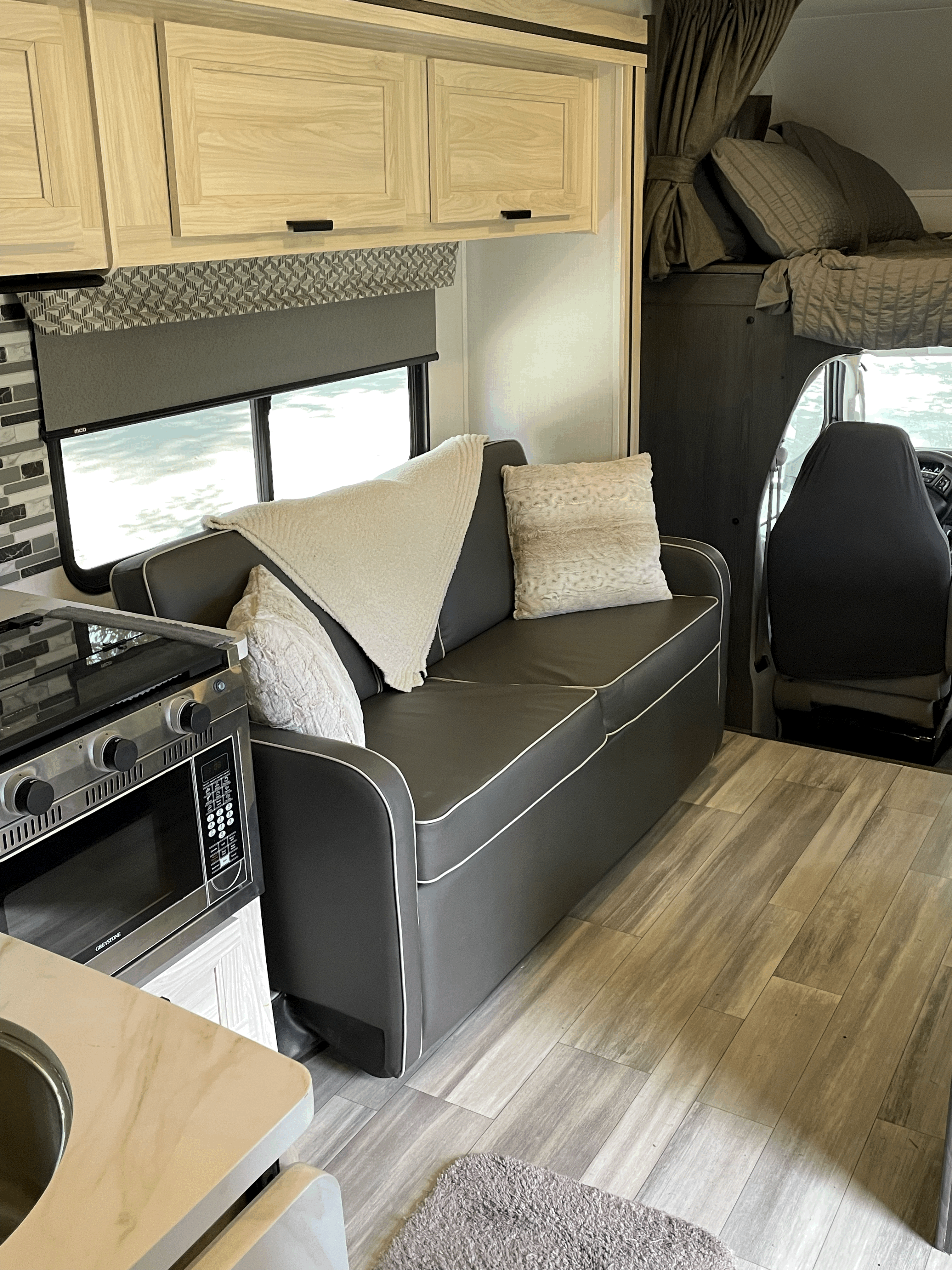 Modern RV rental living space: sofa, compact kitchen with stove/microwave, overhead bed, and wood floors.. Forester LE 2023