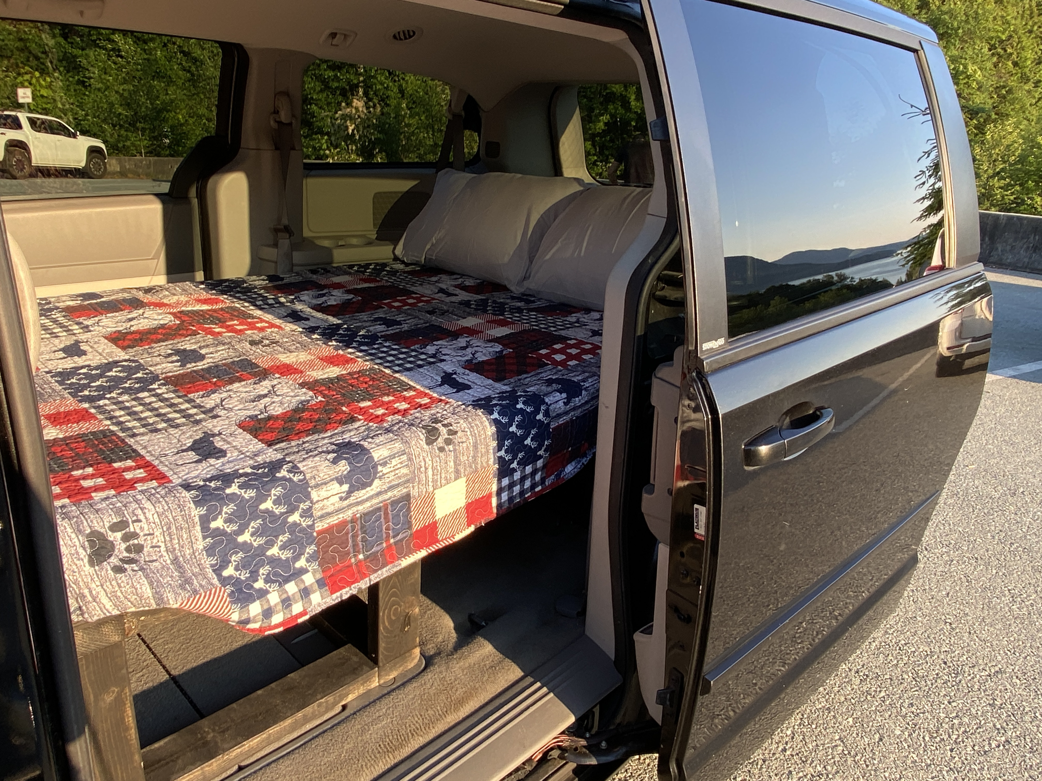 Inside a minivan, a custom bed with patriotic quilt faces an open door with a scenic lake view, ideal for an RV rental.. Dodge Other 2010