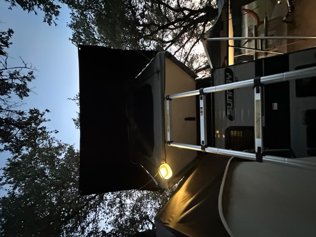 RV rental travel trailer with rooftop tent, illuminated by an outdoor light, and ladder for easy camping access.. Sunset Park RV SunRay 2023