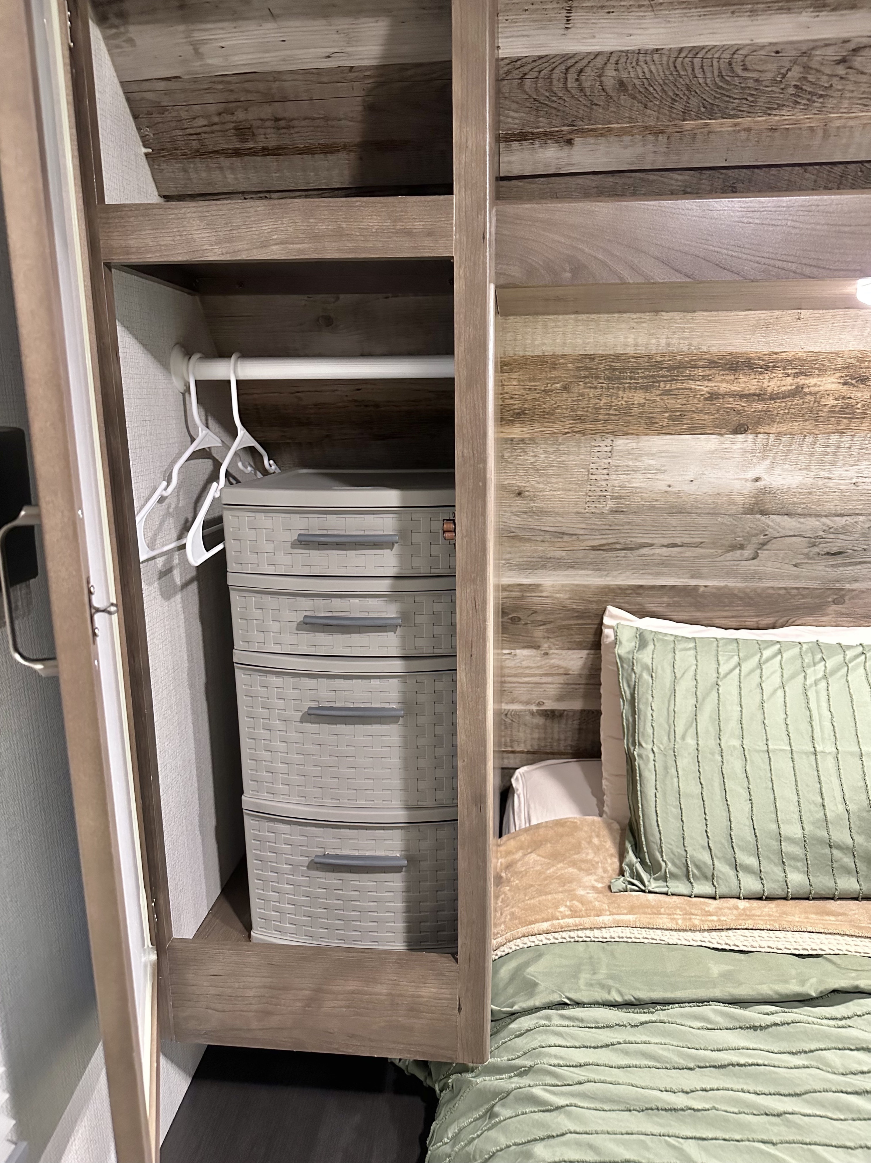 Organized RV rental bedroom: closet with four grey storage drawers & hangers, adjacent to a bed with green bedding and wood-look walls.. Keystone RV Passport SL 2021