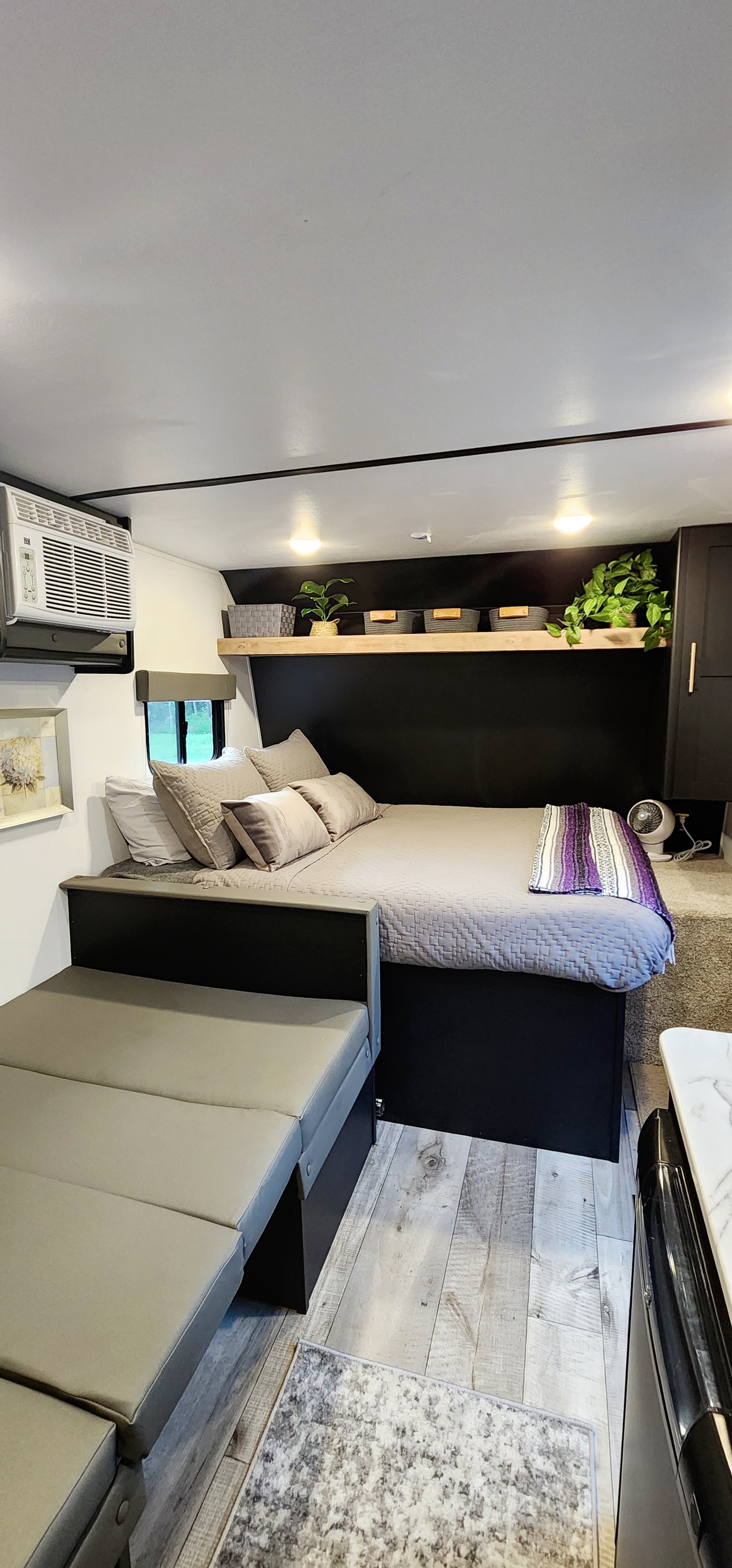 Inside an RV rental, a compact bedroom/living area. Features a bed with pillows, a grey bench, wooden floor, and a shelf with plants.. Keystone RV Springdale 2022
