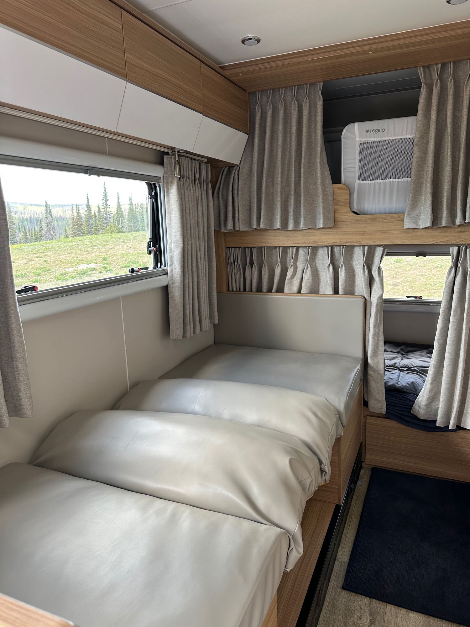 Inside an **rv rental**: two beds, upper bunk with a "Regalo" baby cot, curtains, storage, and a scenic forest view outside.. Black Series Black Series Trailer 2022