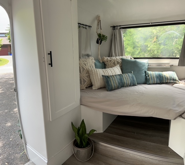 Cozy RV rental interior featuring a bed with pillows, window view of trees, and a plant by the entrance.. Trillium Rv Boler 13' 1975