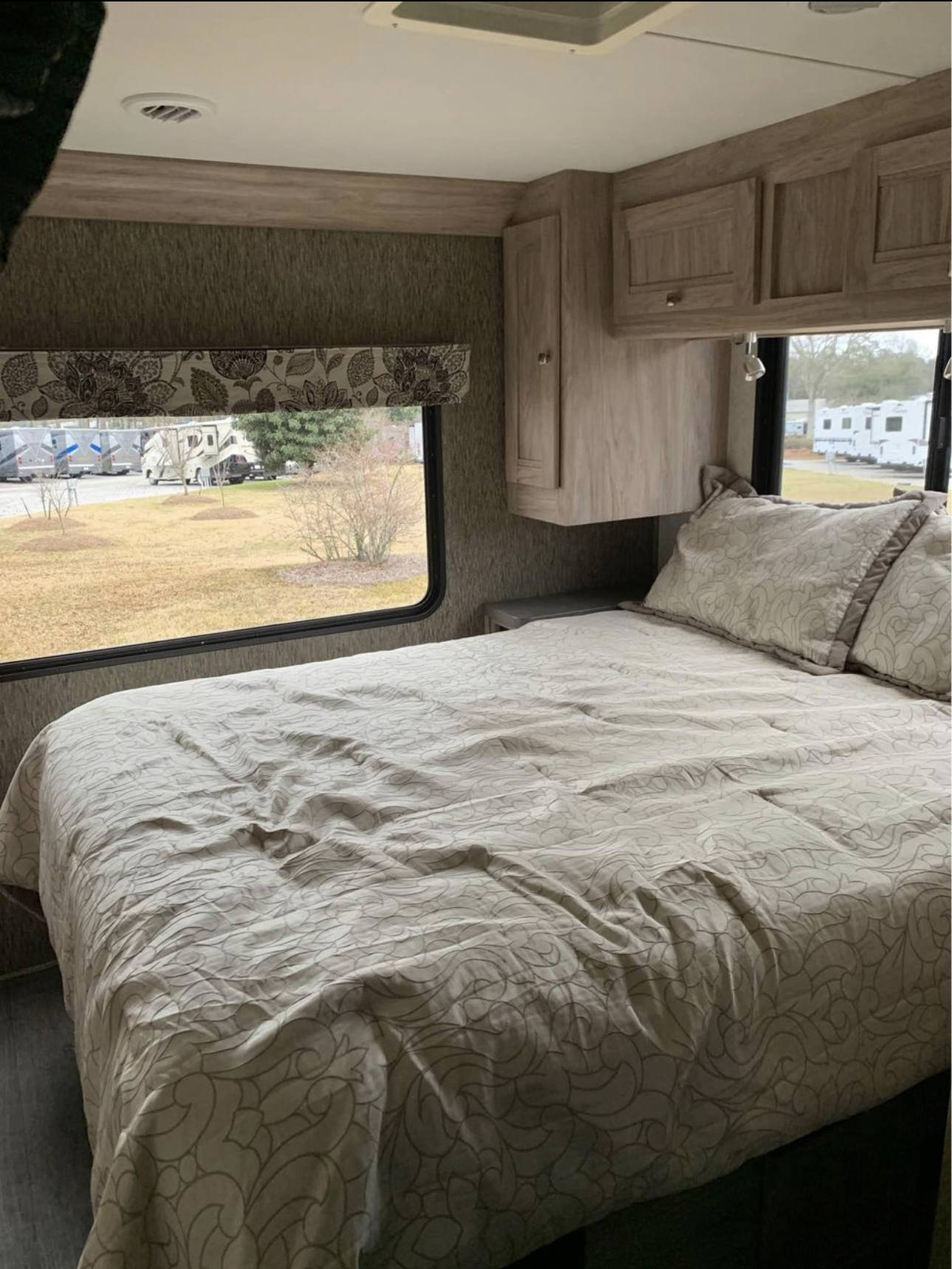 An RV rental's bedroom, featuring a comfy bed with patterned linens, overhead cabinets, and a window view of an RV park.. Forester LE 2018